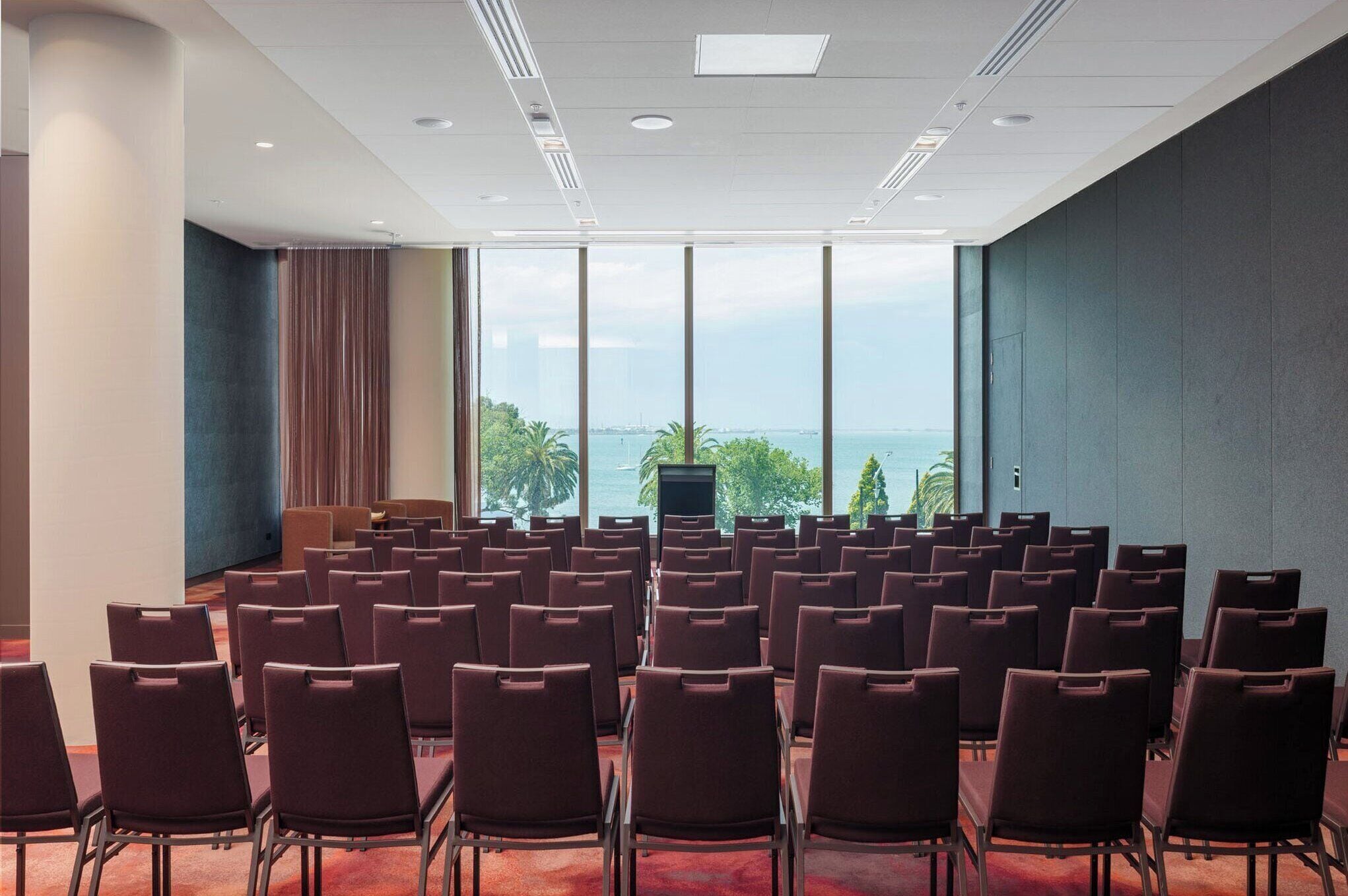 A conference room with rows of chairs, large windows with a view of the sea, and a podium.
