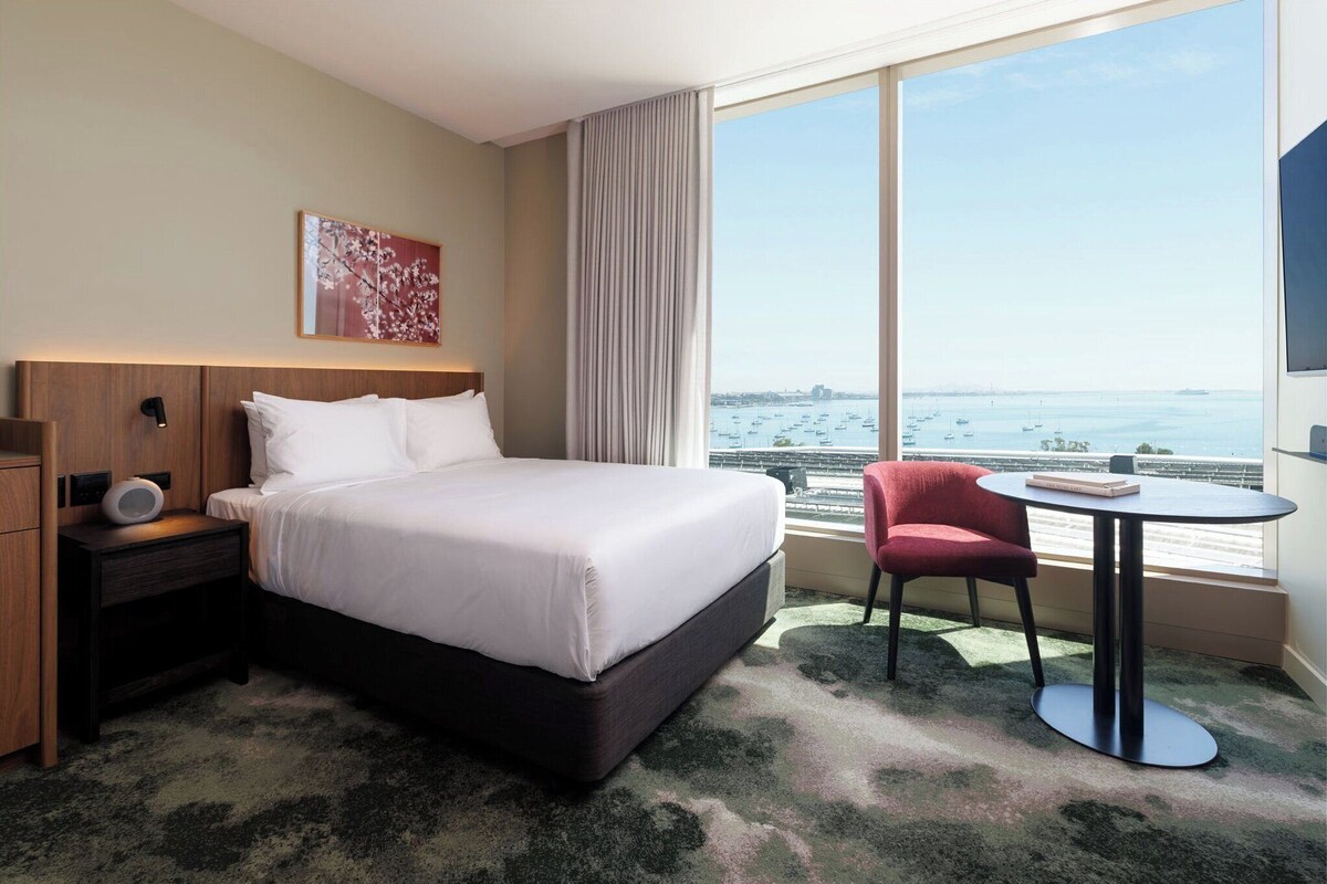 A hotel room with a large bed, a bedside table with a lamp, a chair, a small table, and a large window with a view of the sea and boats.