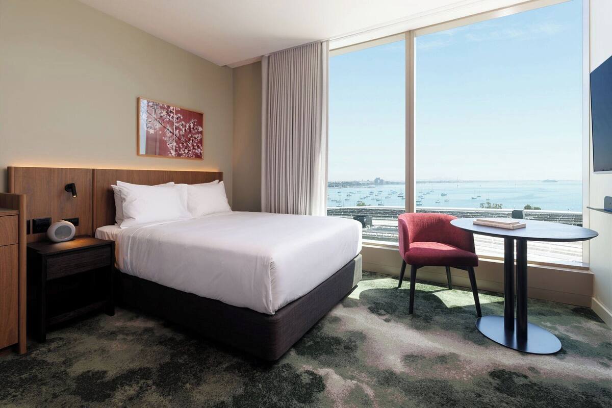 Une chambre d’hôtel avec un grand lit, une table de chevet avec une lampe, une chaise, une petite table et une grande fenêtre donnant sur la mer et les bateaux.