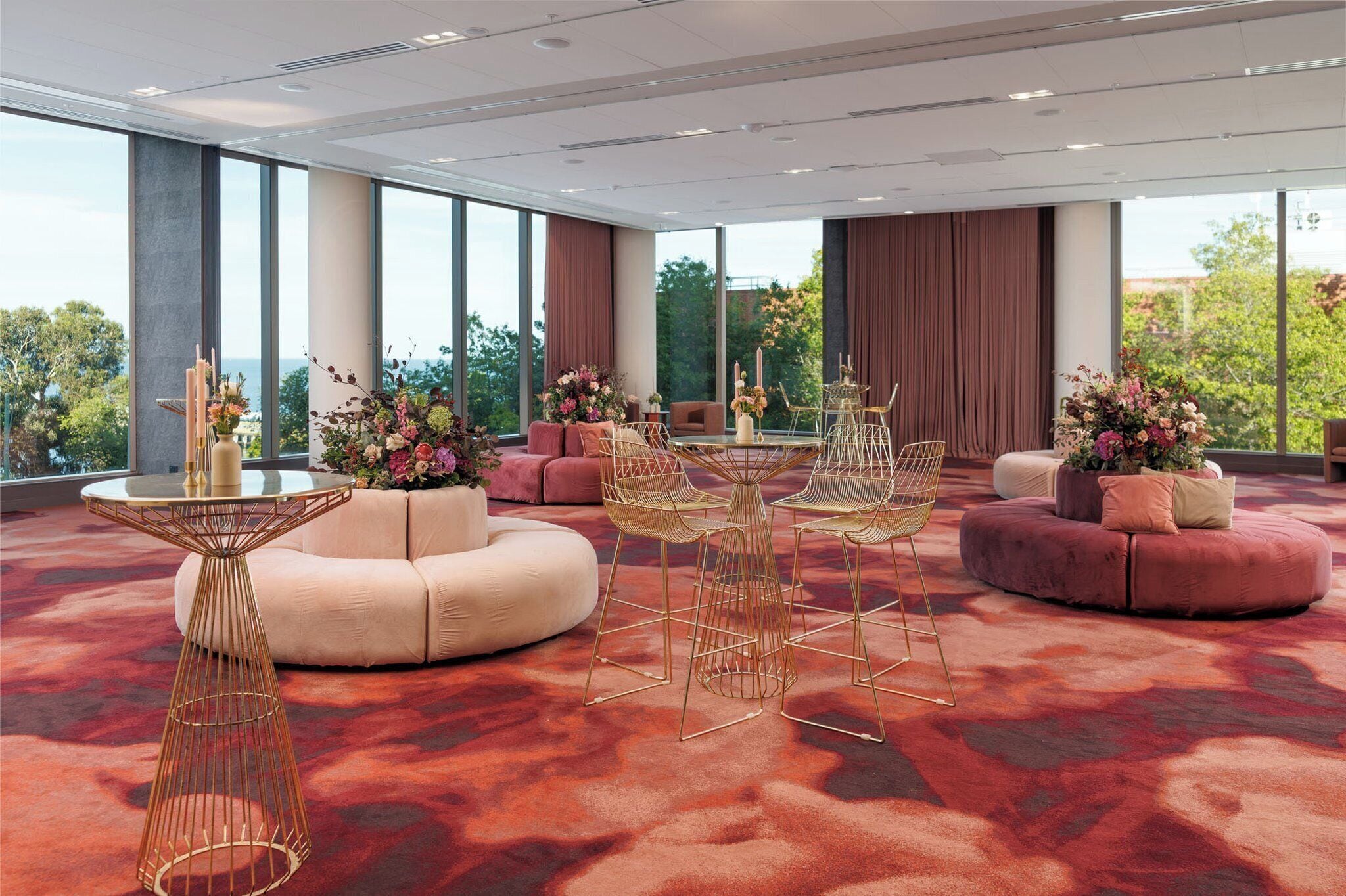 Una habitación de hotel moderna con ventanas amplias, alfombra roja, una mesa redonda con sillas y un sofá con una mesita auxiliar.