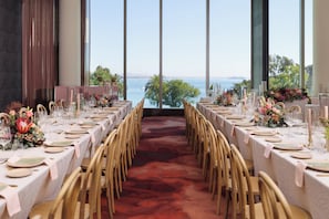 Une longue table entourée de chaises, une moquette rouge et une vue sur la mer par de grandes fenêtres.