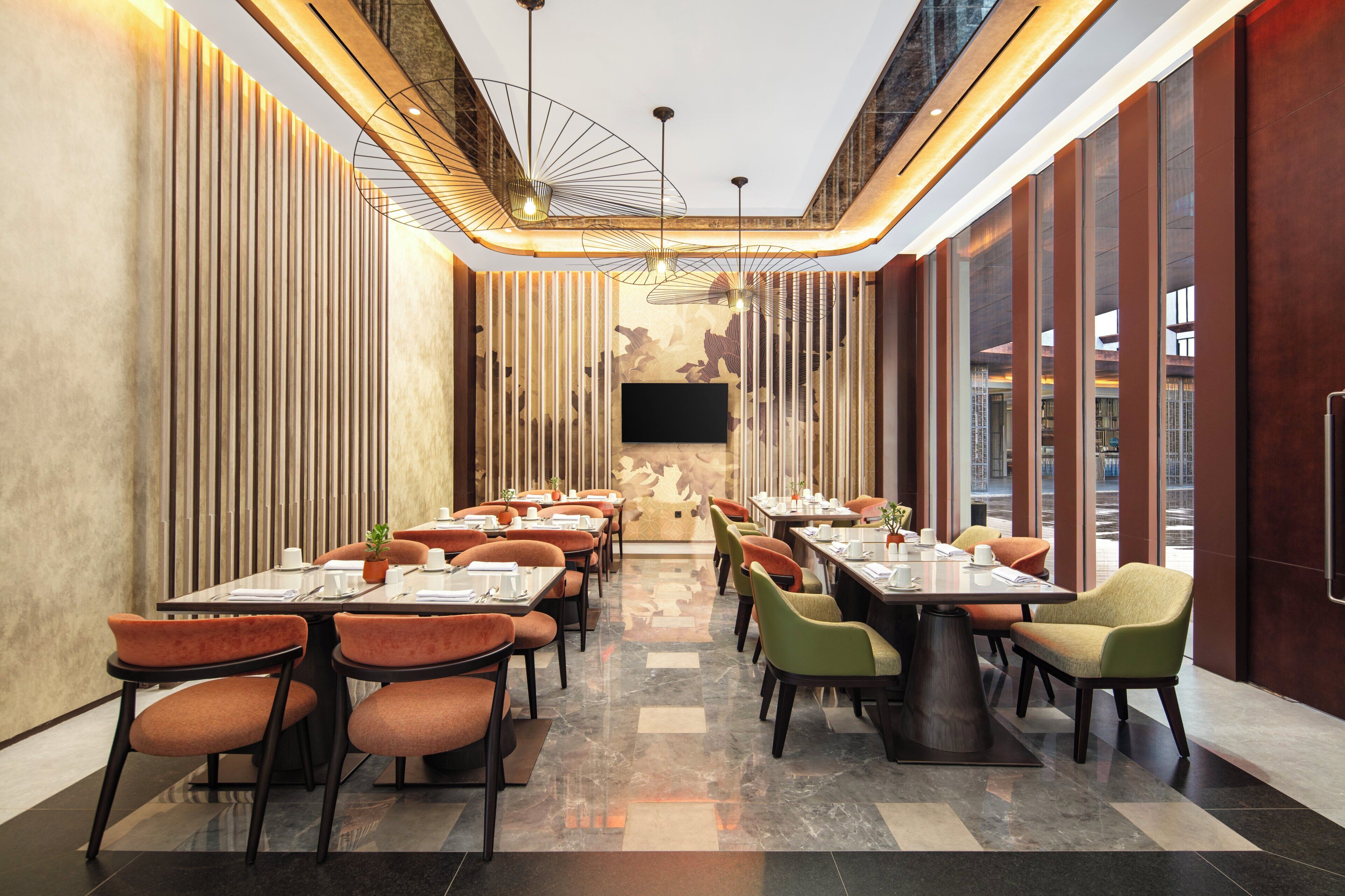 A modern restaurant with a long table layout, high ceilings, and a large TV screen.