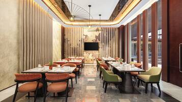 A modern restaurant with a long table layout, high ceilings, and a large TV screen.