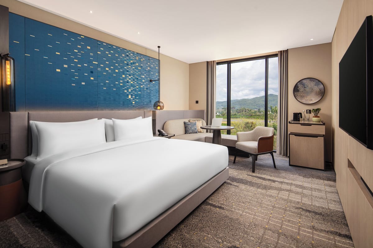Quarto de hotel moderno com uma cama grande, área de estar com uma mesa, televisão e vista para o exterior.