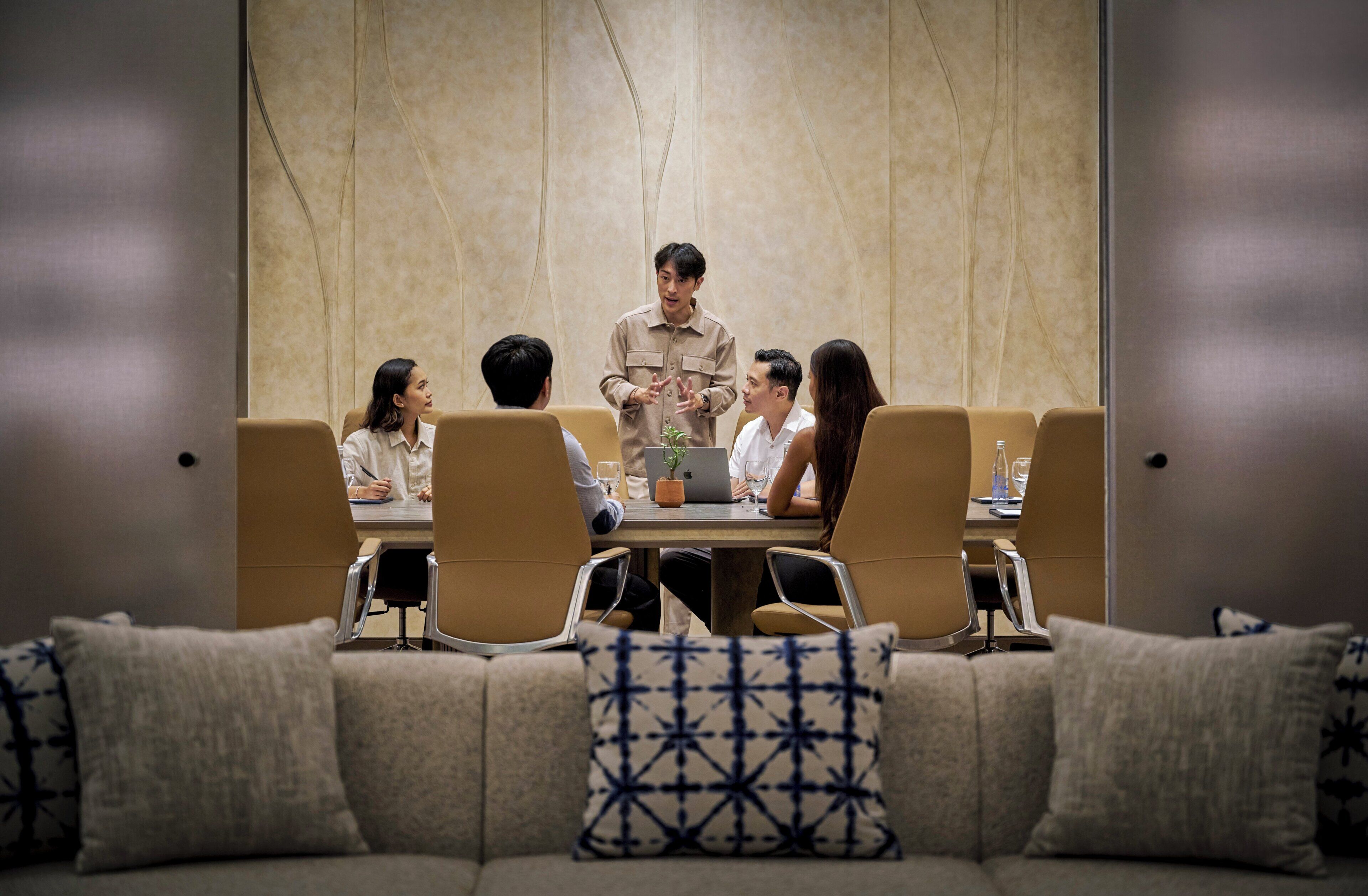 A meeting room with a table, chairs, and a person presenting.