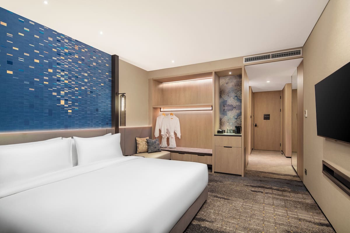 Quarto de hotel moderno com uma cama grande, área de estar, TV e banheiro visível por uma porta aberta.