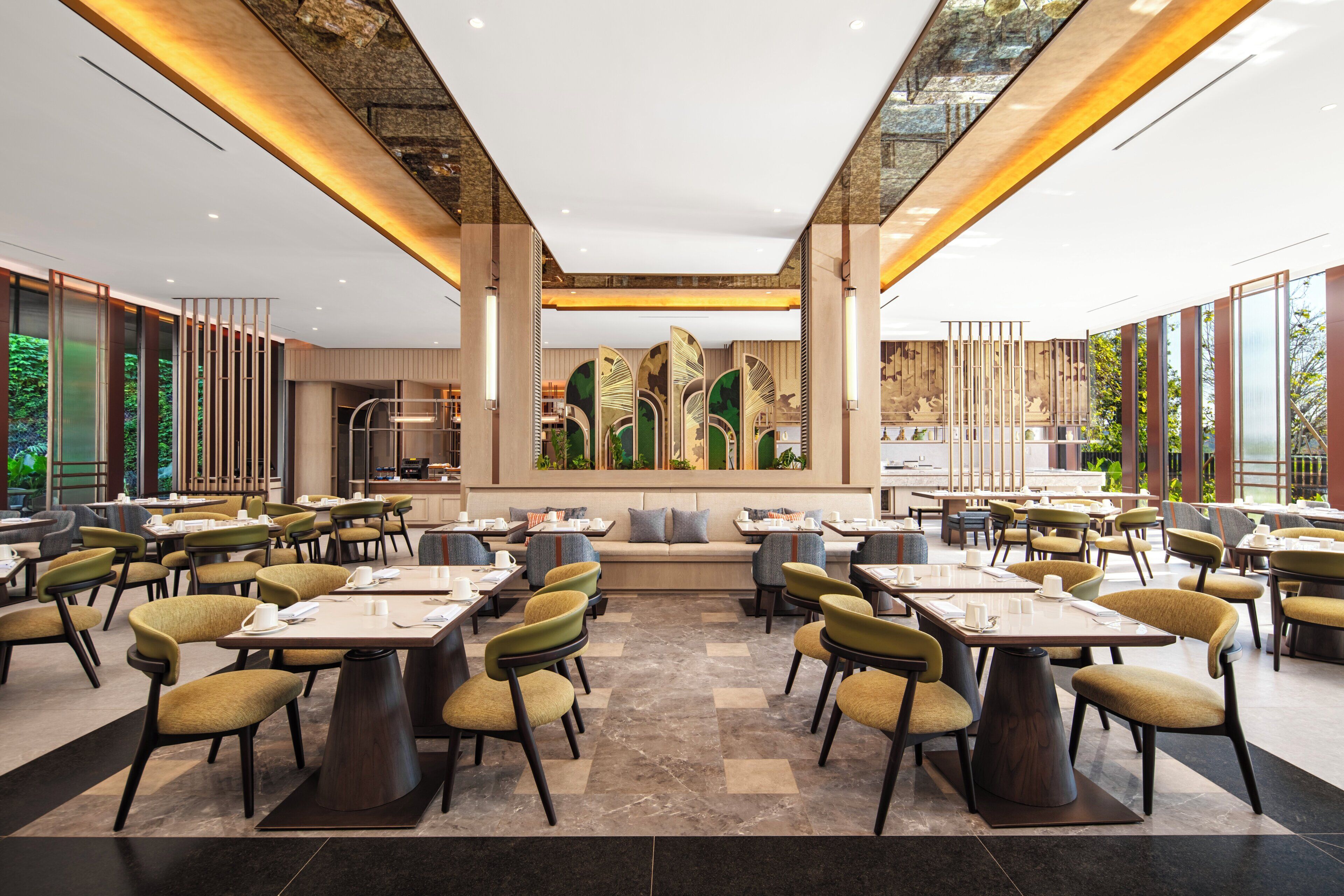 A spacious dining area with multiple tables and chairs, a central seating area, and large windows offering a view of greenery.