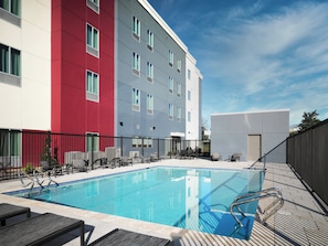 Una piscina con sillas de descanso, una valla y un edificio de fachadas rojas y grises.