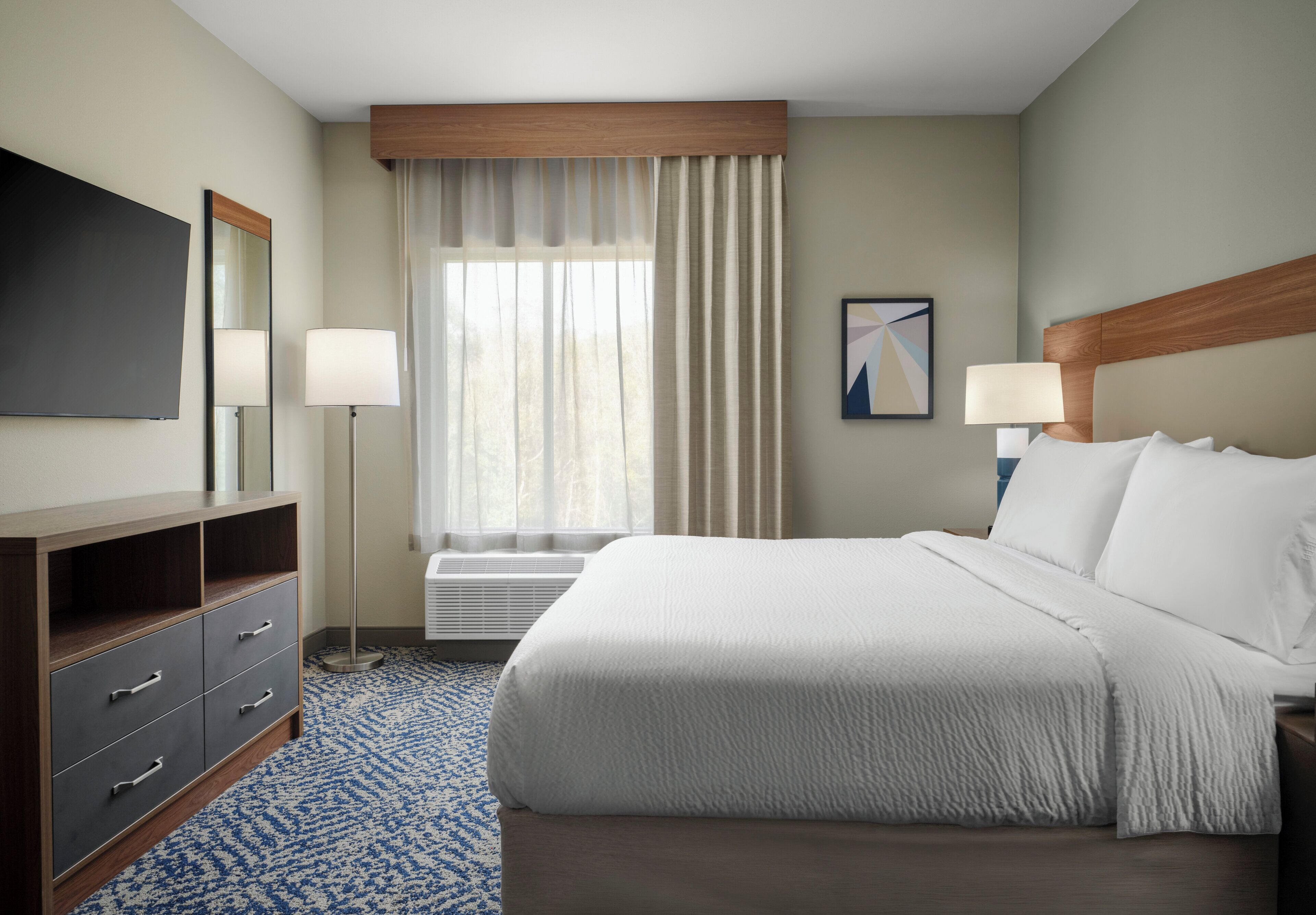Quarto de hotel com cama, escrivaninha, cadeira, televisão de tela plana, luminária, janela com cortinas e um quadro na parede.