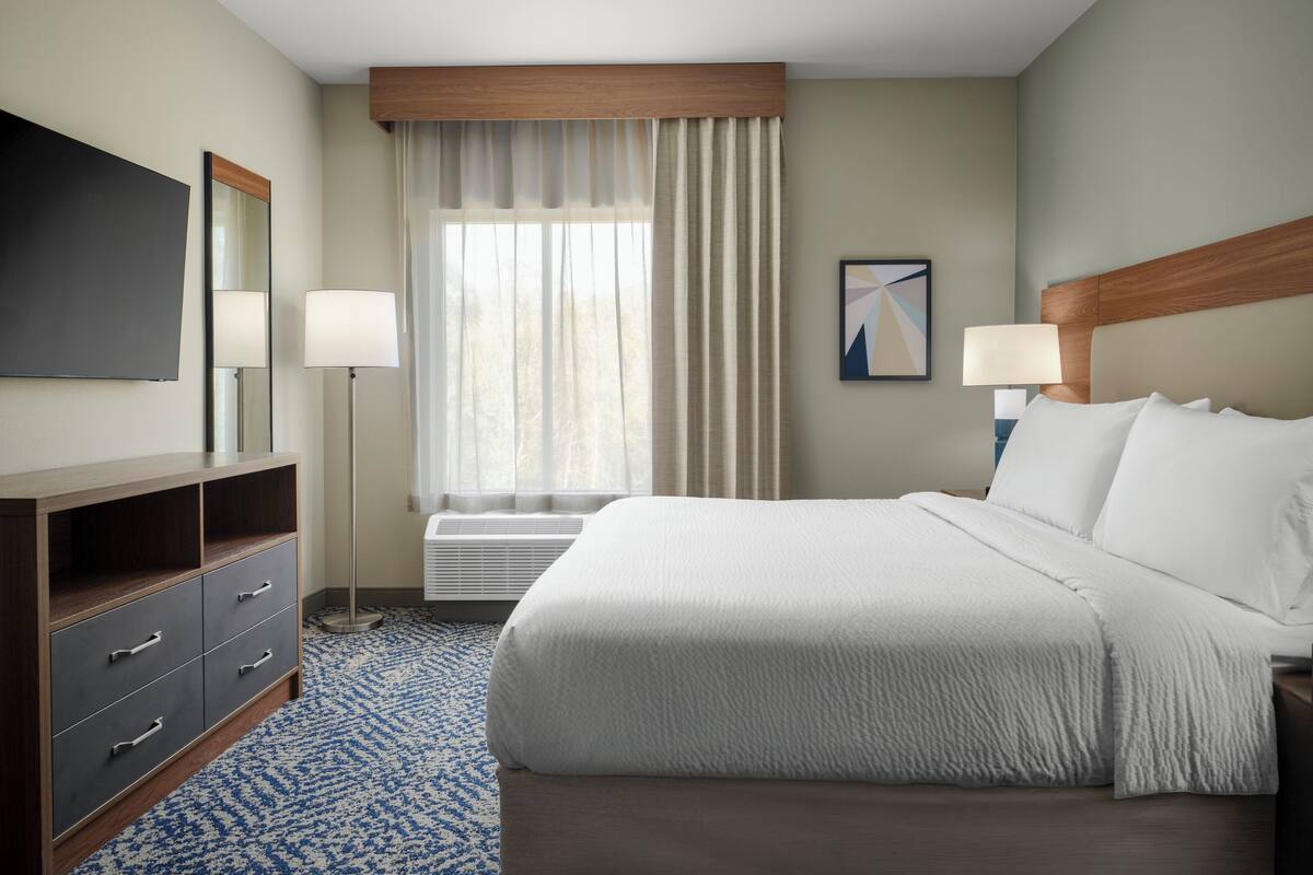 A hotel room with a bed, a desk, a chair, a flat-screen TV, a lamp, a window with curtains, and a painting on the wall.