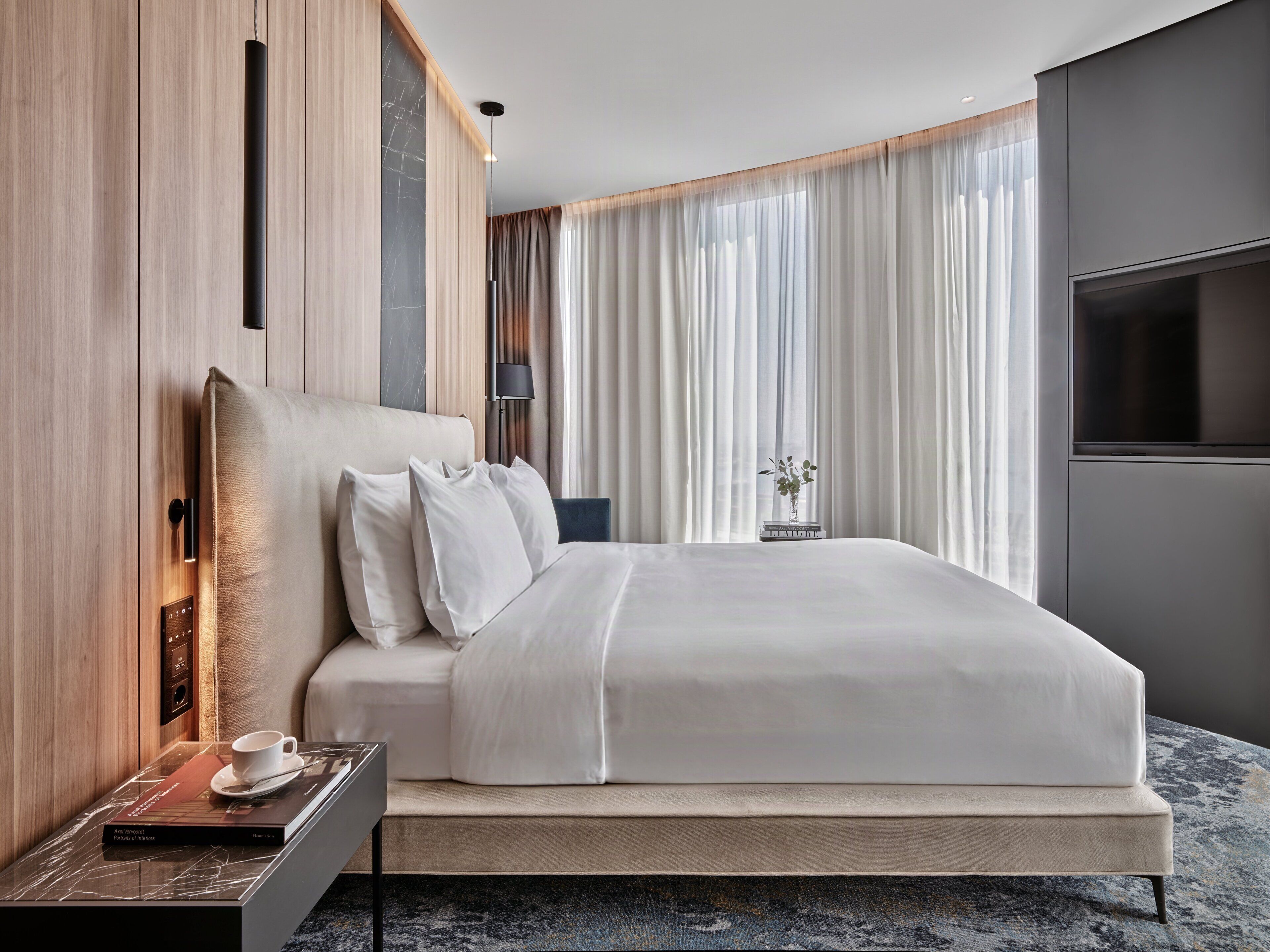A modern hotel room with a large bed, a bedside table with a book and a cup, a TV, and a window with sheer curtains.