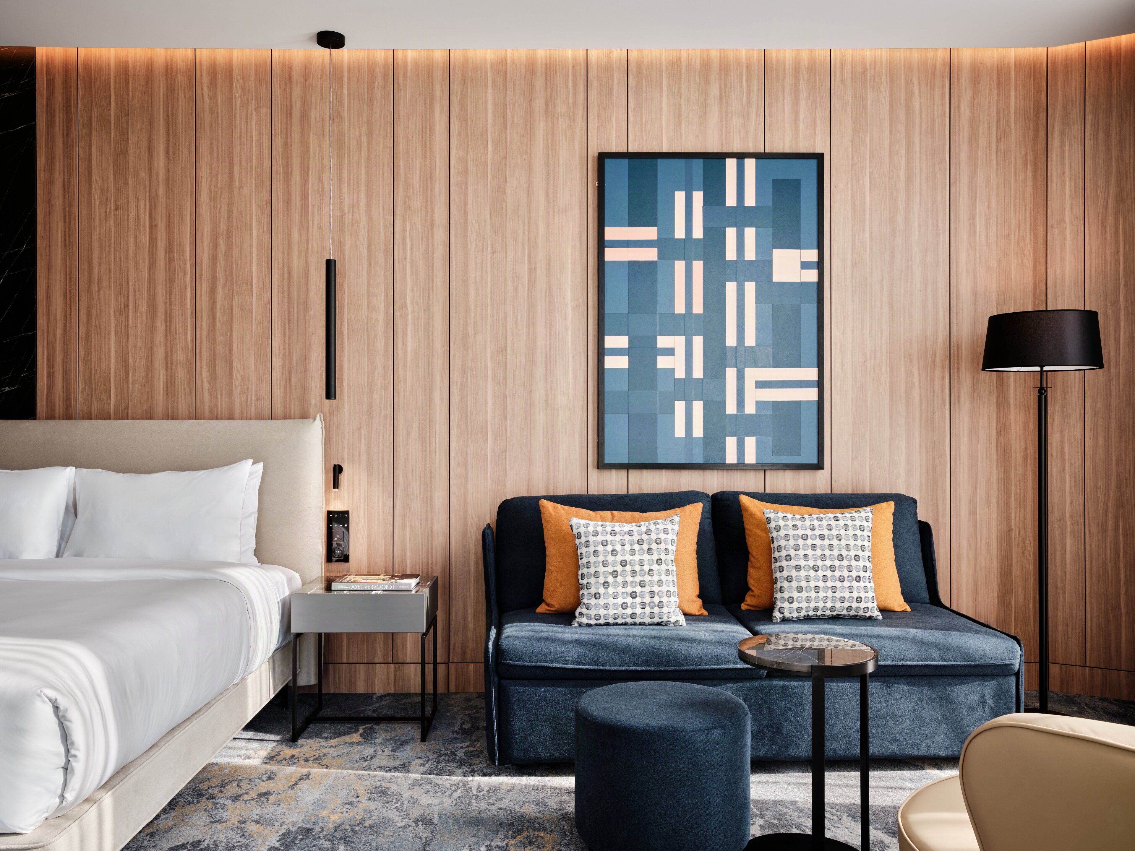 A modern hotel room with a bed, a sofa, a small table, a lamp, and a wall-mounted artwork.