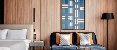 A modern hotel room with a bed, a sofa, a small table, a lamp, and a wall-mounted artwork.