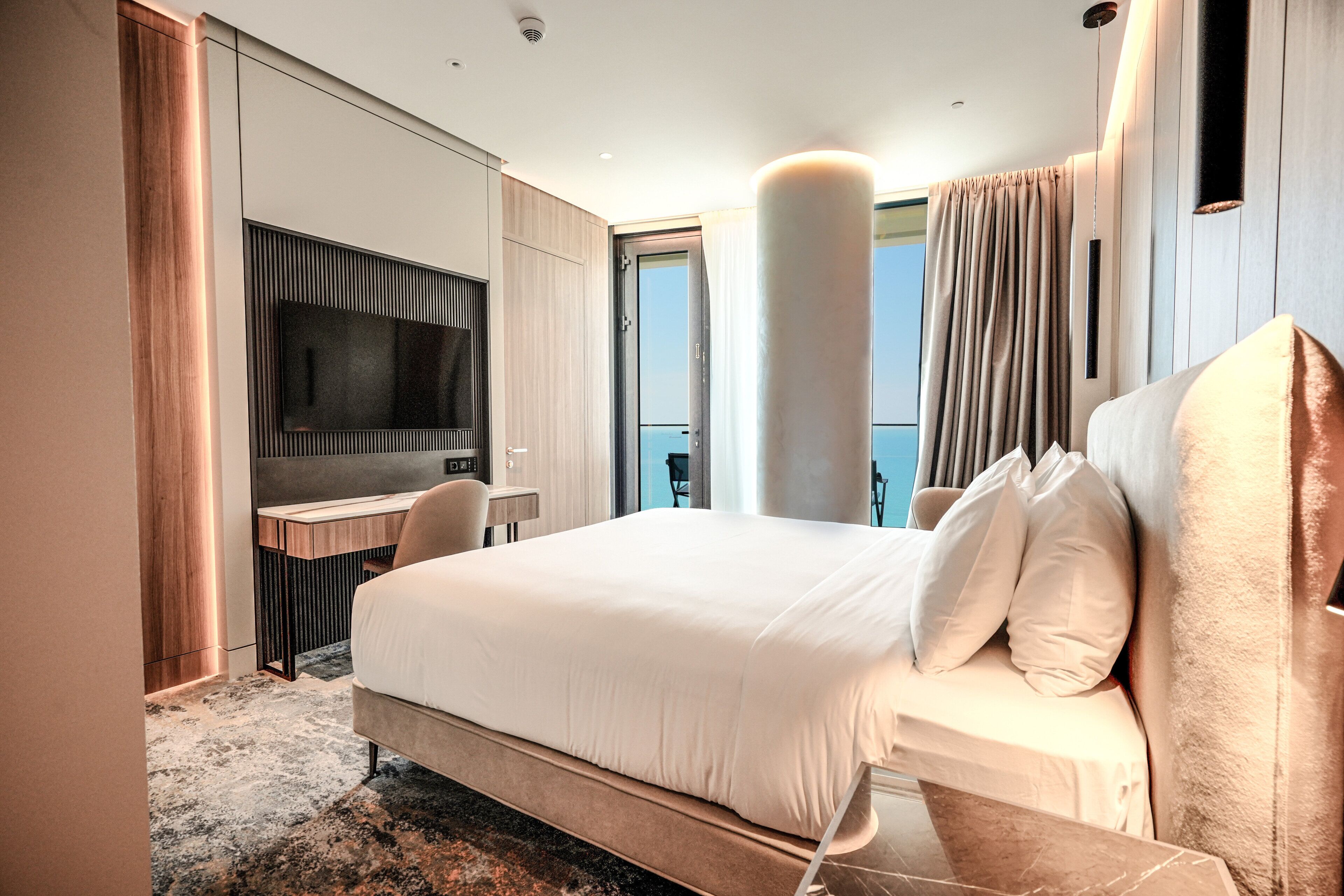 A modern hotel room with a large bed, a desk, a flat-screen TV, and a balcony with curtains.