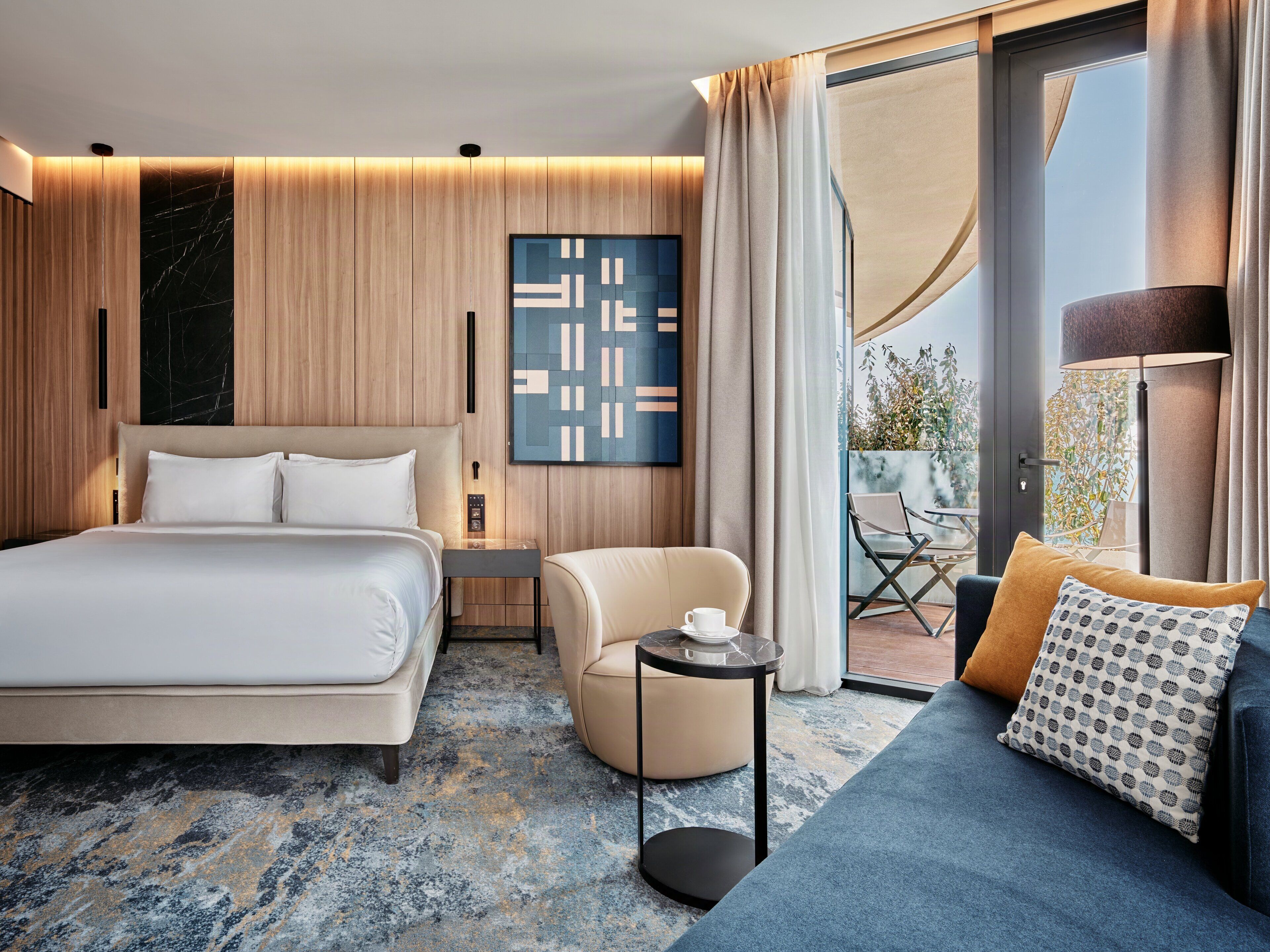 A modern hotel room with a bed, a sofa, a small table, and a balcony with outdoor furniture.