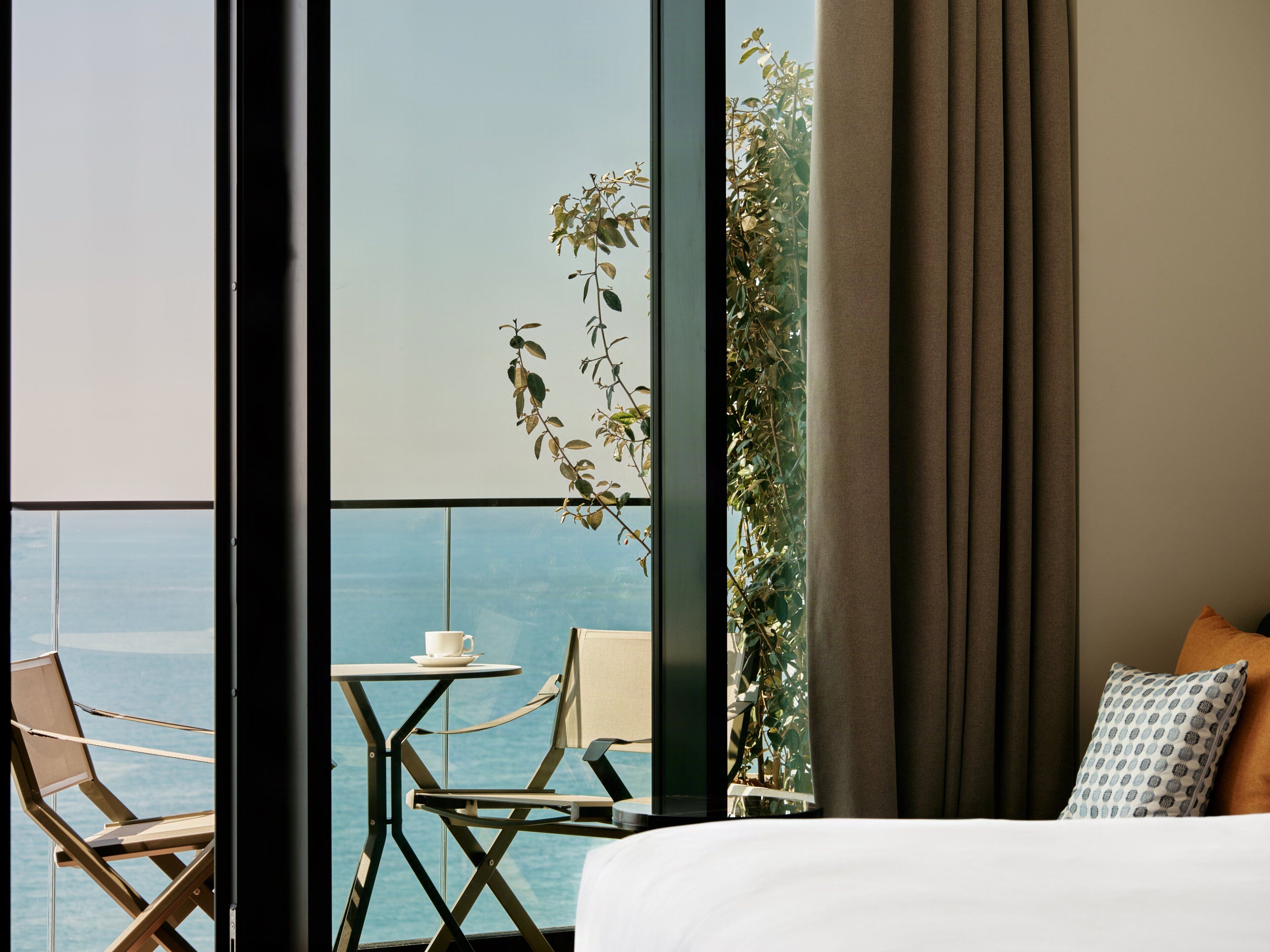 A hotel room with a bed, a small table with a cup, two chairs, and a view of the sea through large windows.