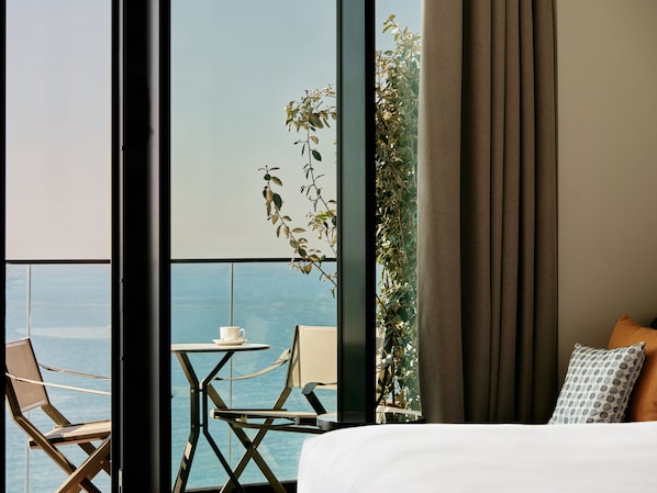 Habitación de hotel con cama, una mesita con una taza, dos sillas y vista al mar a través de amplias ventanas.