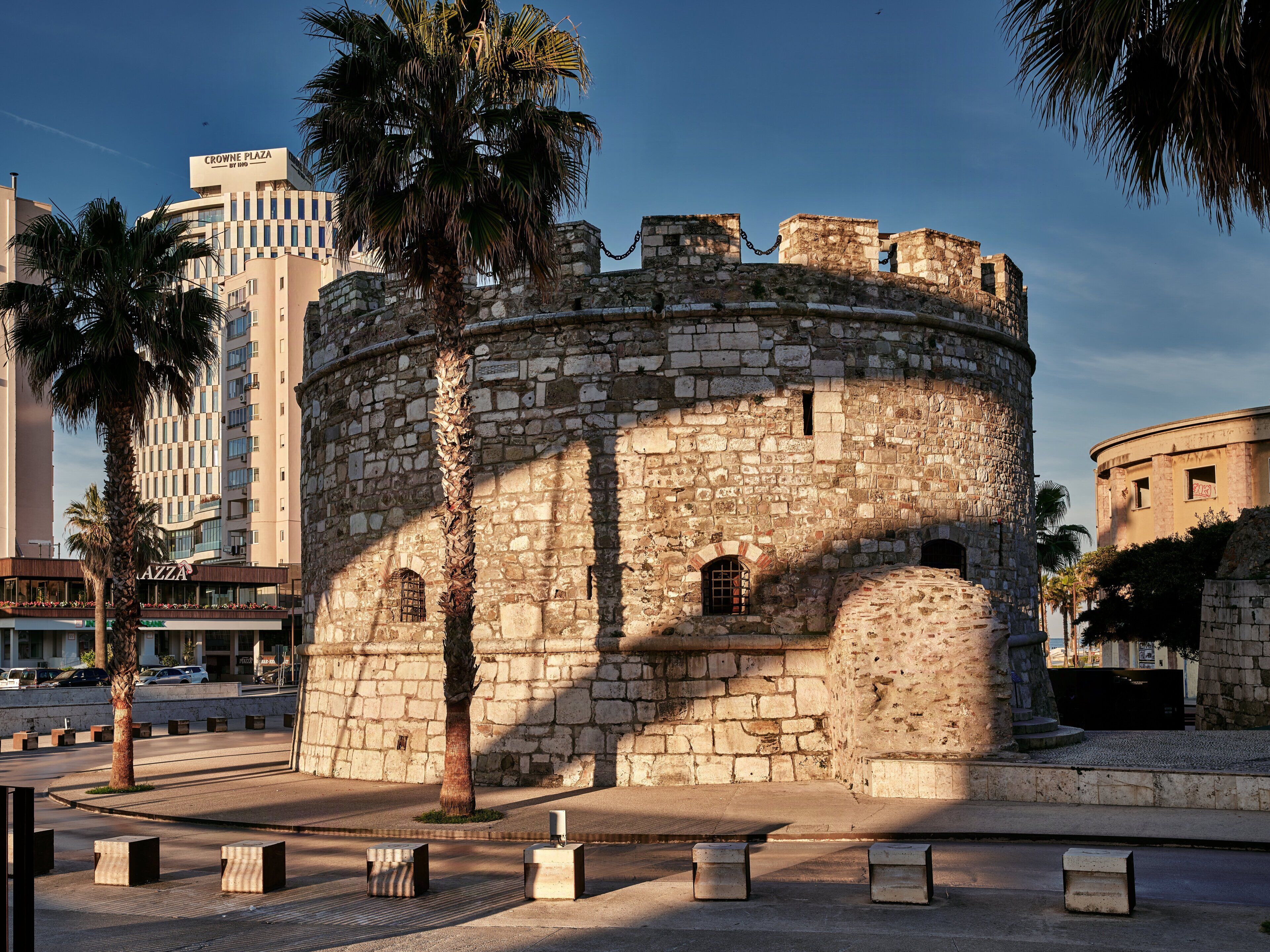 A historic stone tower with a circular base, surrounded by modern buildings and palm trees.