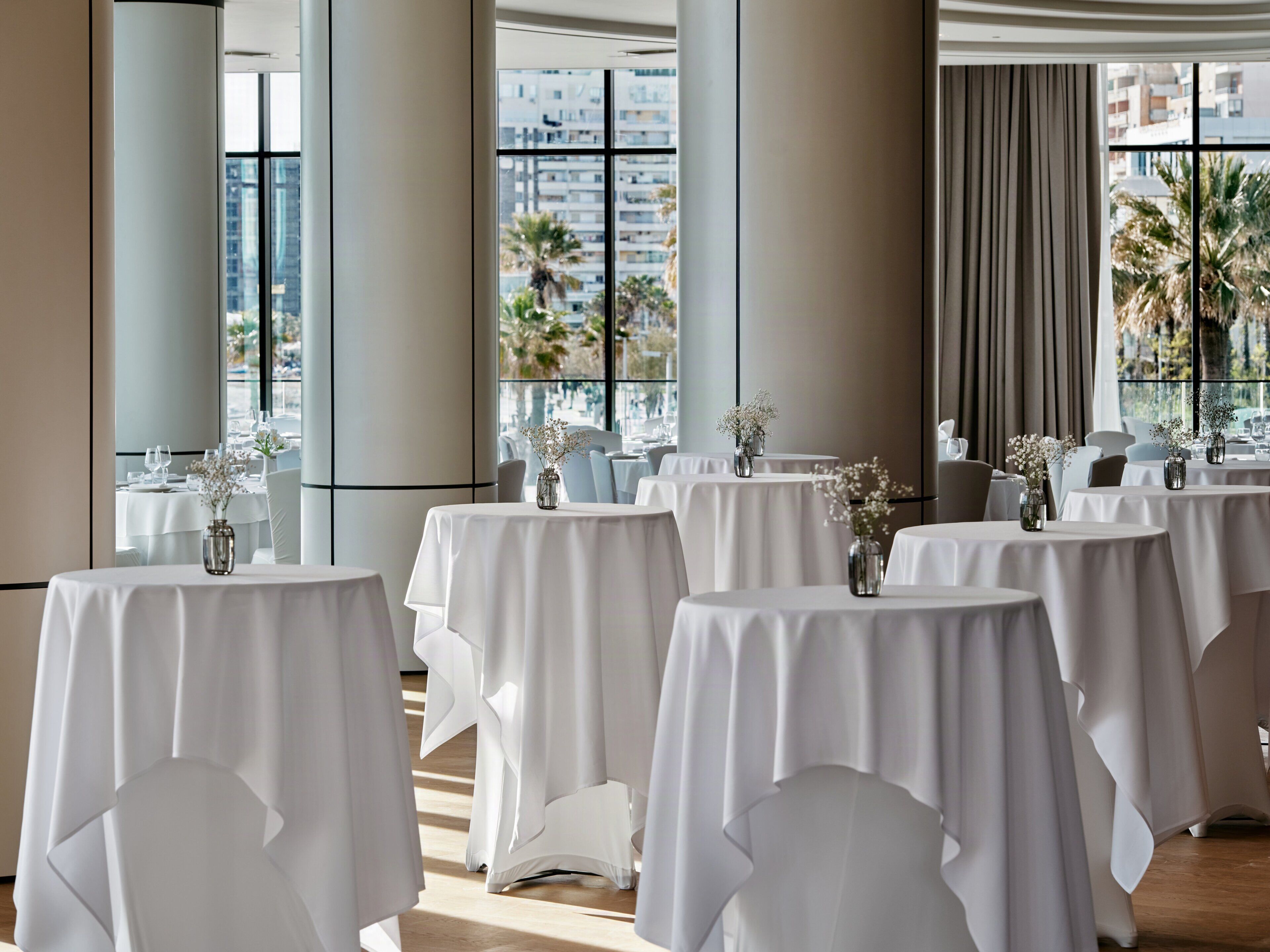 A hotel dining area with round tables, white tablecloths, and vases of flowers.