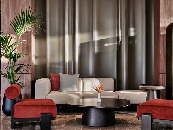 A modern living room with a sofa, red ottoman, coffee table, and a potted plant.