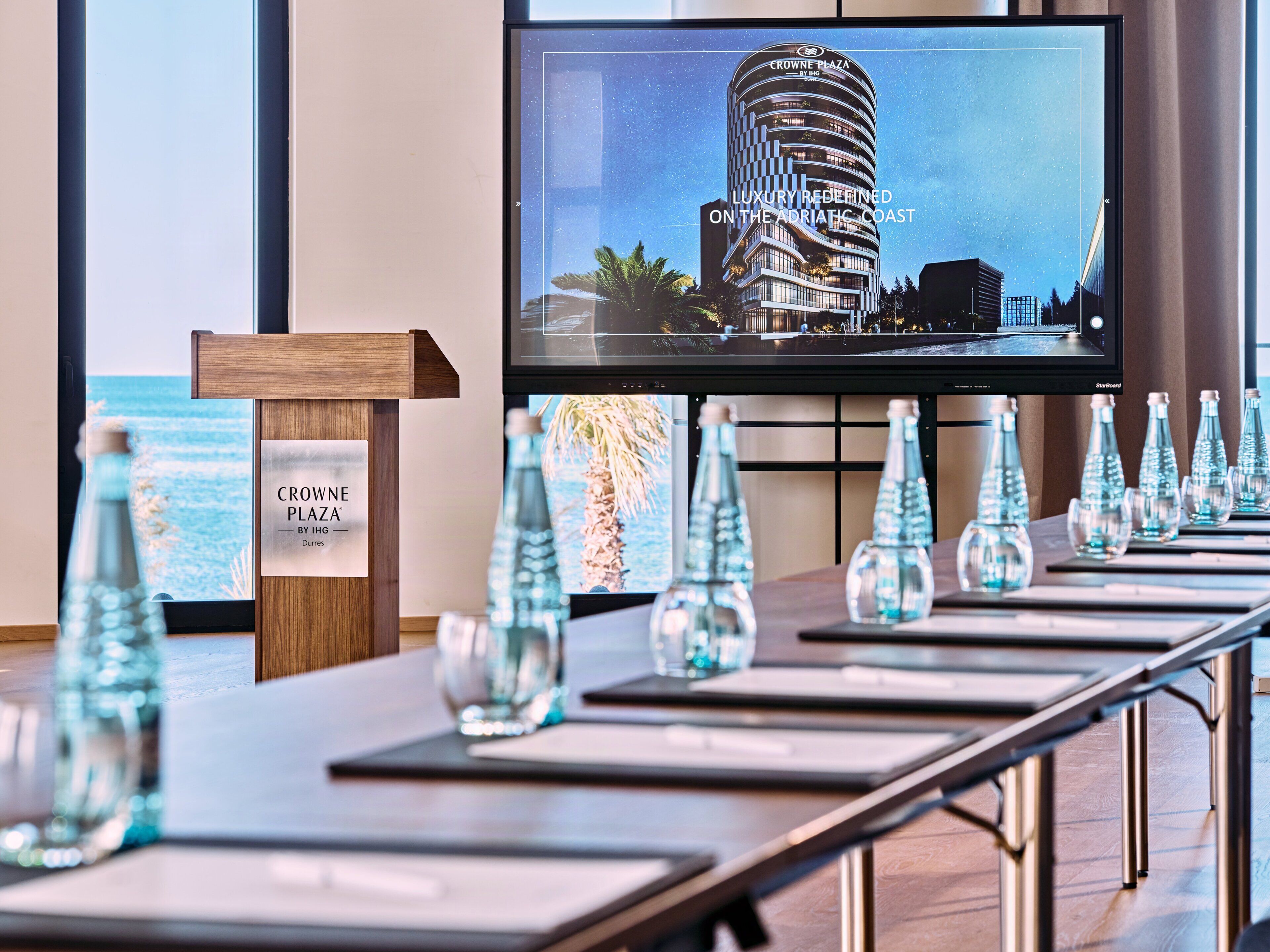 Een vergaderruimte met een groot scherm waarop een gebouw van 'Crowne Plaza' staat afgebeeld en een bord met de vermelding 'by IHG'.