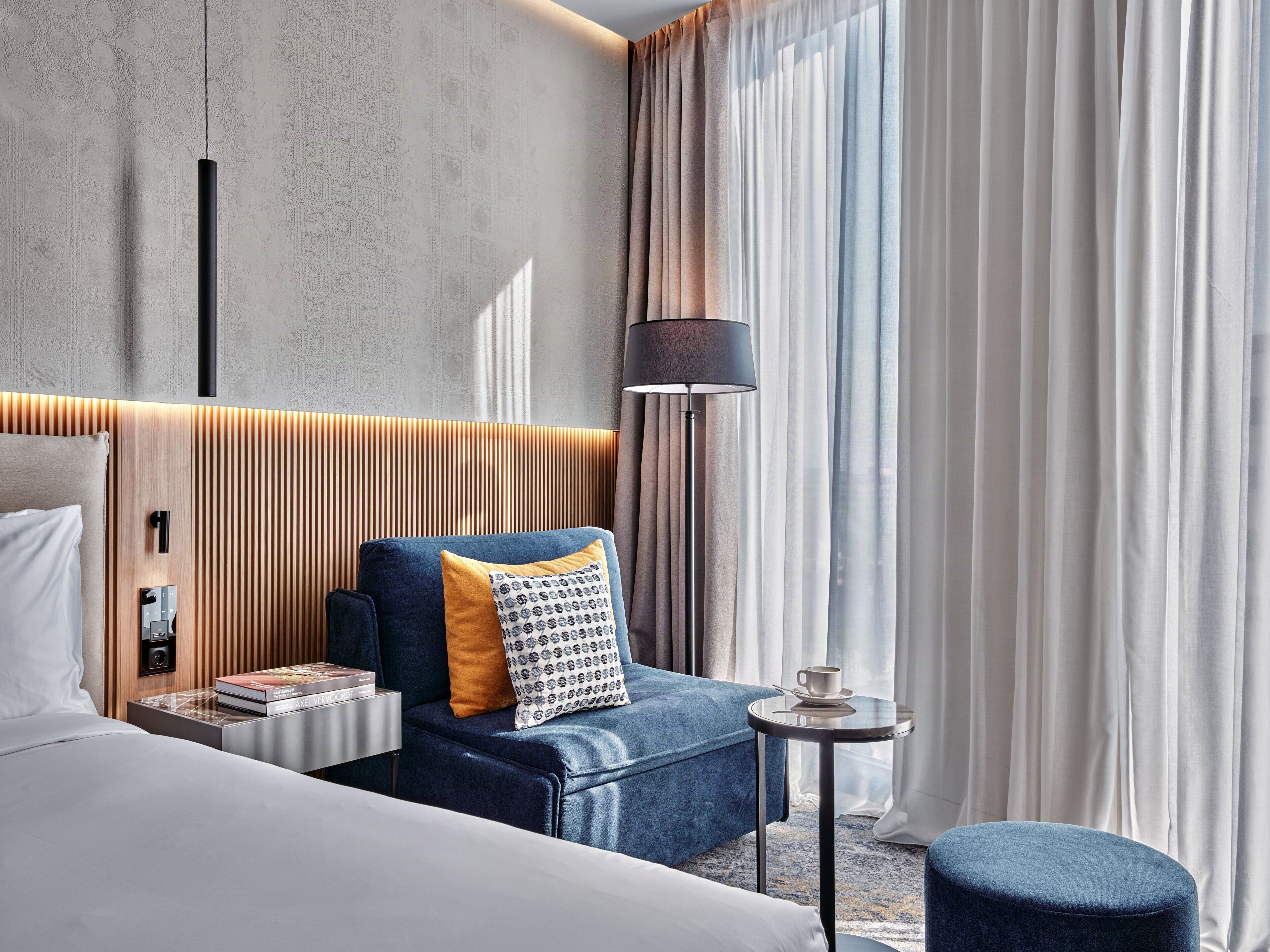 Een moderne hotelkamer met een bed, een zithoek met een bank en fauteuil, een nachtkastje met een lamp, en uitzicht op het landschap door dunne gordijnen.