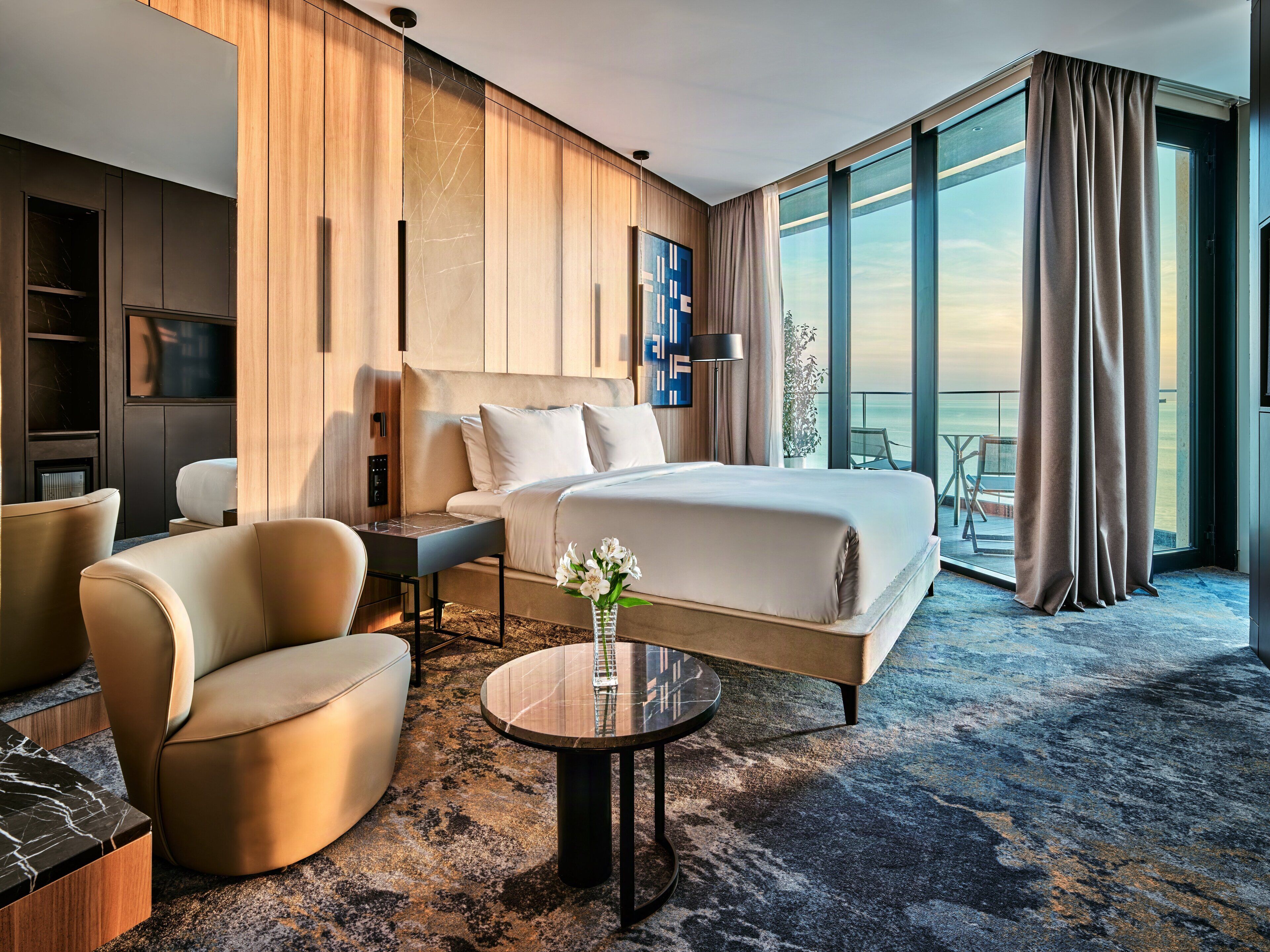 Una habitación de hotel moderna con una cama grande, una zona de estar con una mesita de café, un televisor y un balcón con vistas.