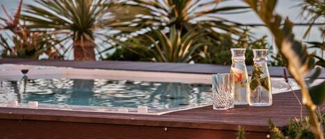 A poolside area with two glass bottles containing beverages, a wooden deck, and a view of the ocean with palm trees in the background.