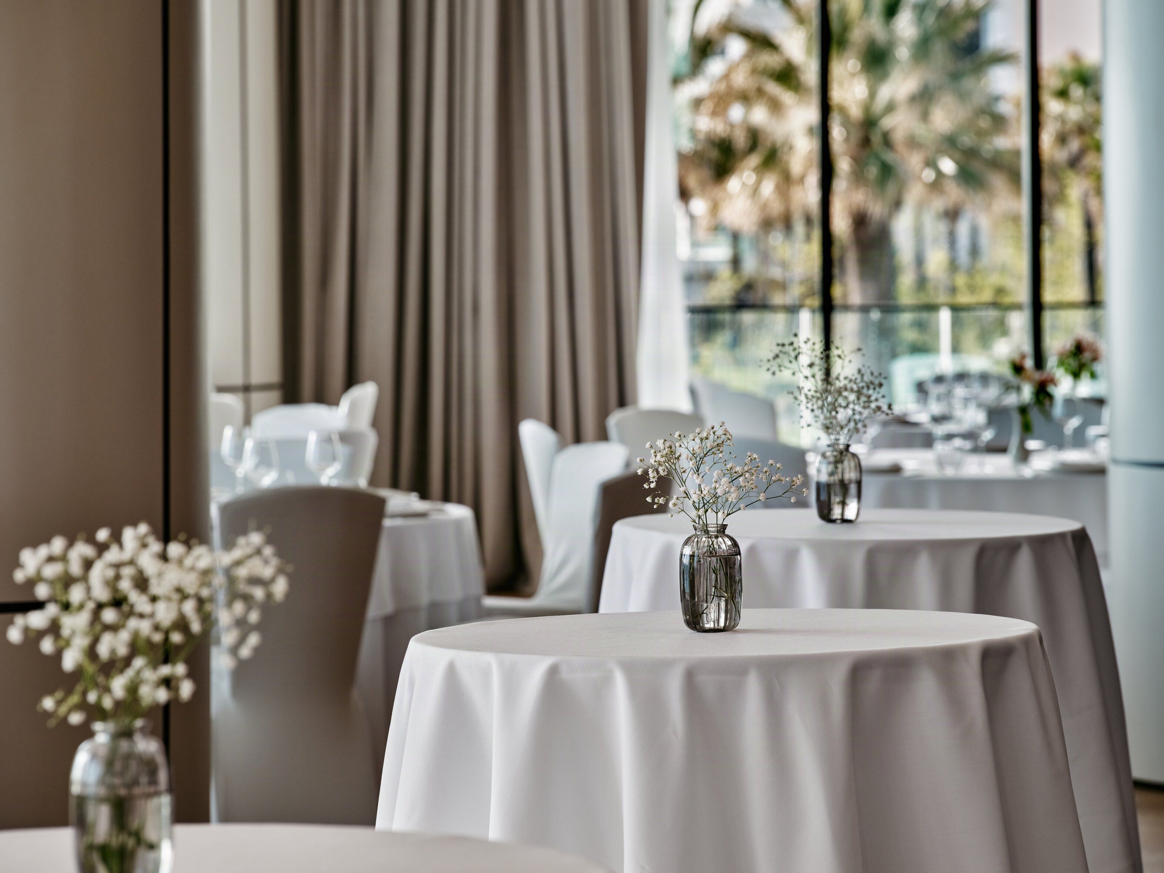 A restaurant with round tables, white tablecloths, and vases with flowers.