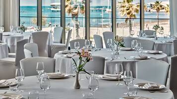 A restaurant with round tables set for dining, large windows with a view of the beach and ocean, and white tablecloths.