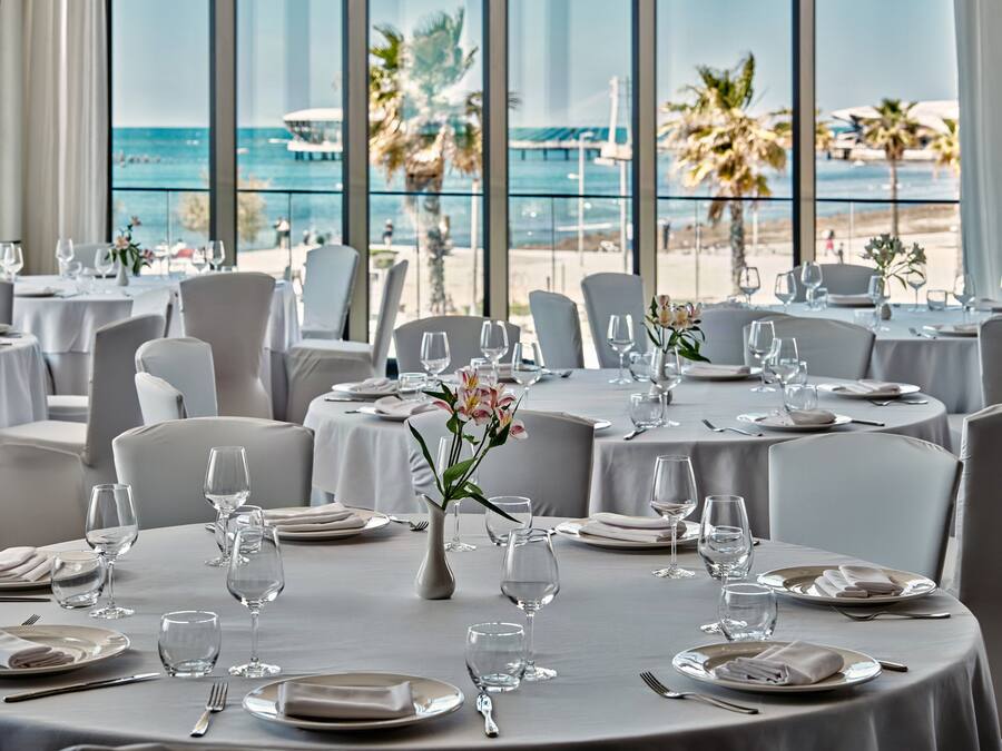 A restaurant with round tables set for dining, large windows with a view of the beach and ocean, and white tablecloths.