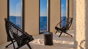 Une pièce dotée de grandes fenêtres, de deux chaises noires, d’une petite table ronde et d’un sol sablonneux.