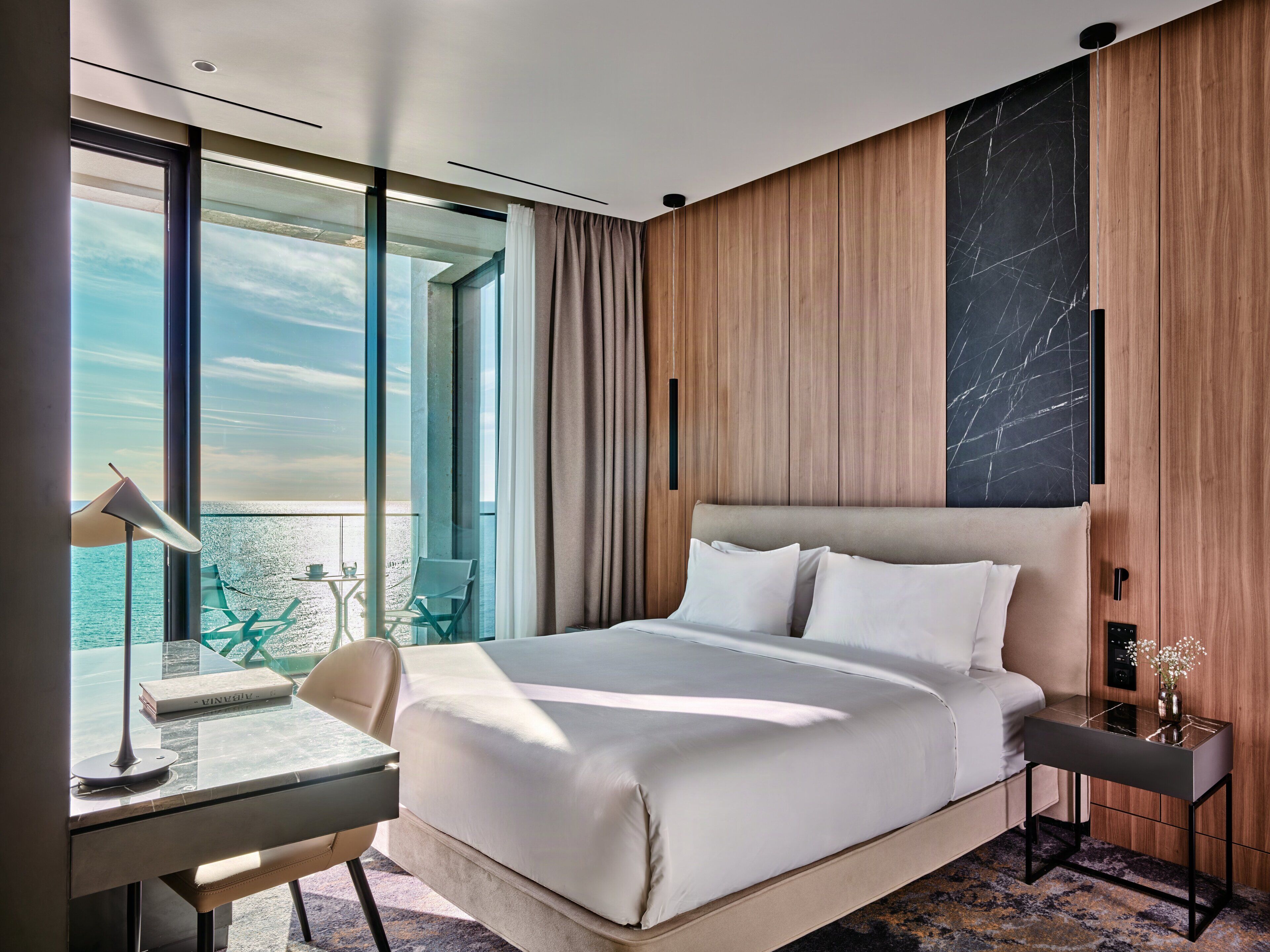 A hotel room with a large bed, a desk with a lamp, a chair, and a view of the sea.