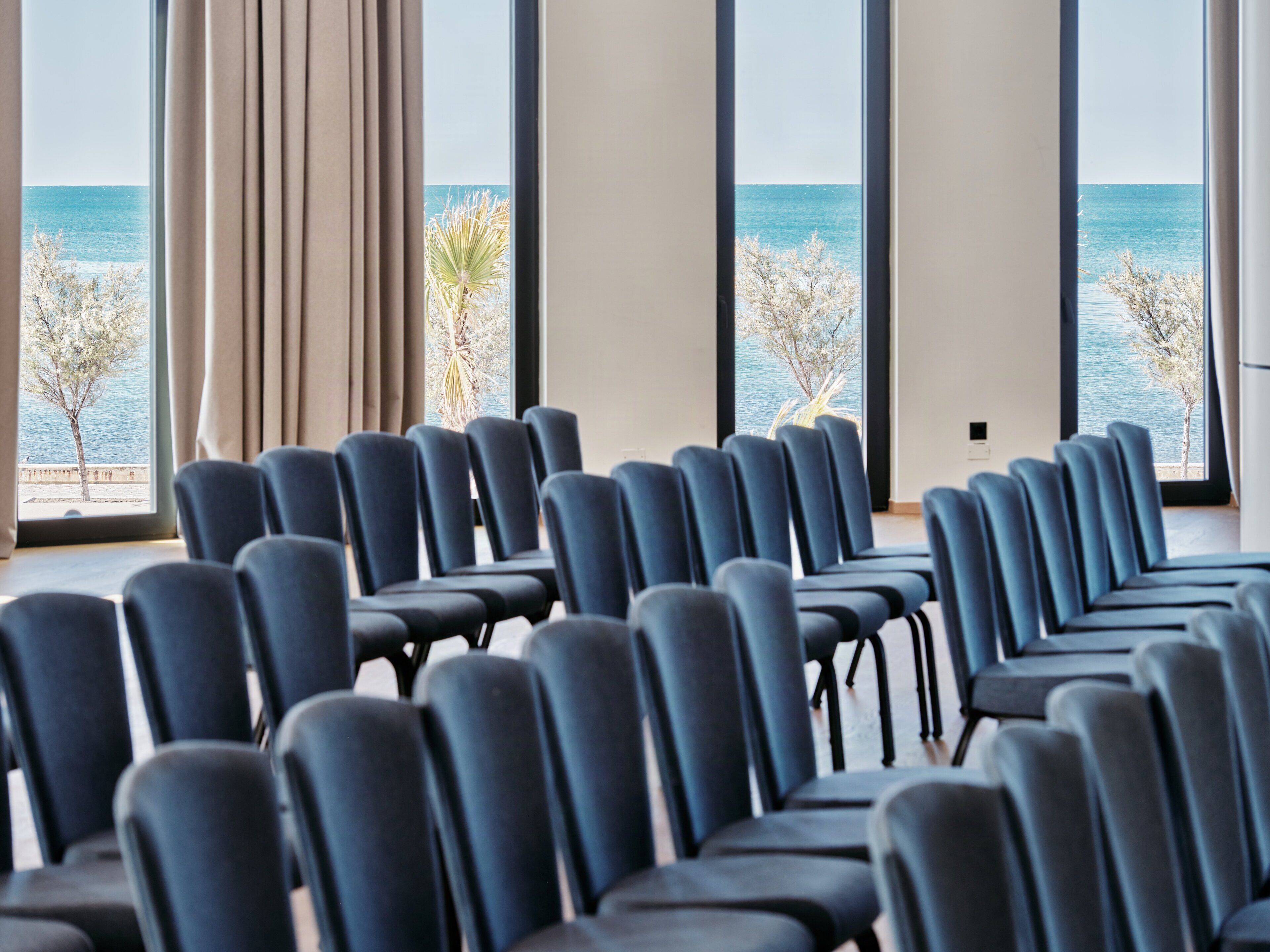 A conference room with rows of chairs facing a large window with a view of the sea and palm trees.