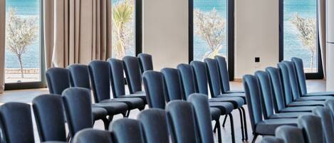 A conference room with rows of chairs facing a large window with a view of the sea and palm trees.