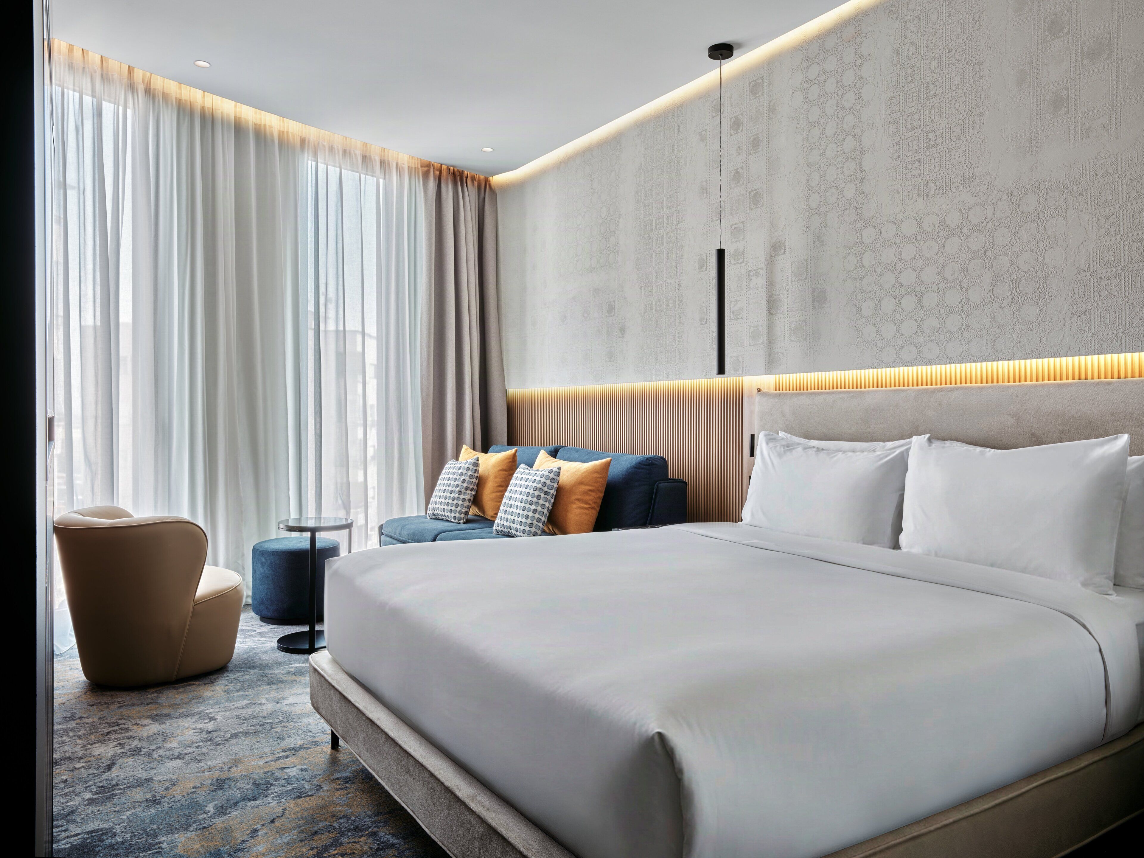 A modern hotel room with a large bed, a sofa, a chair, and a small table.