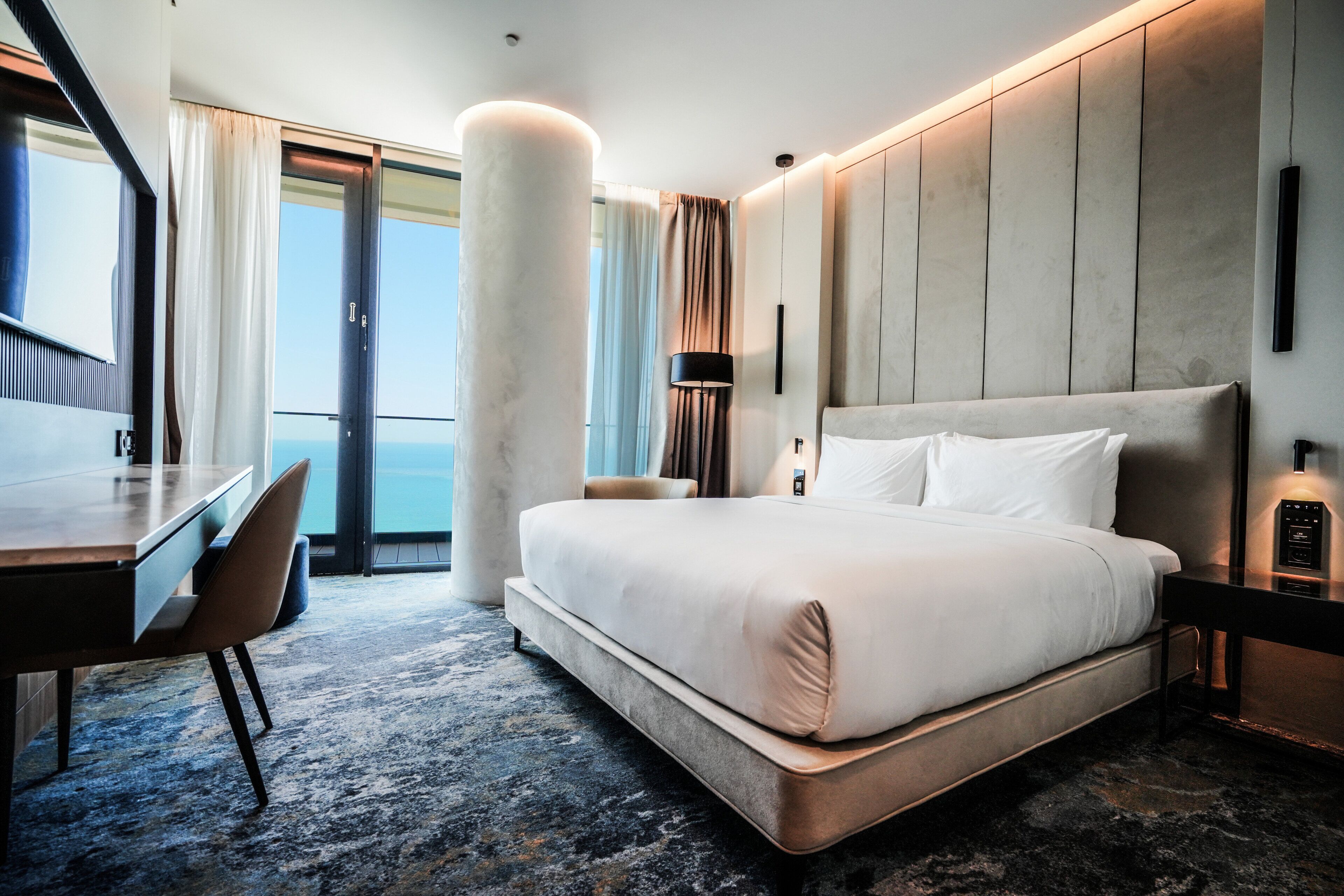 A modern hotel room with a large bed, a desk, and a view of the sea.
