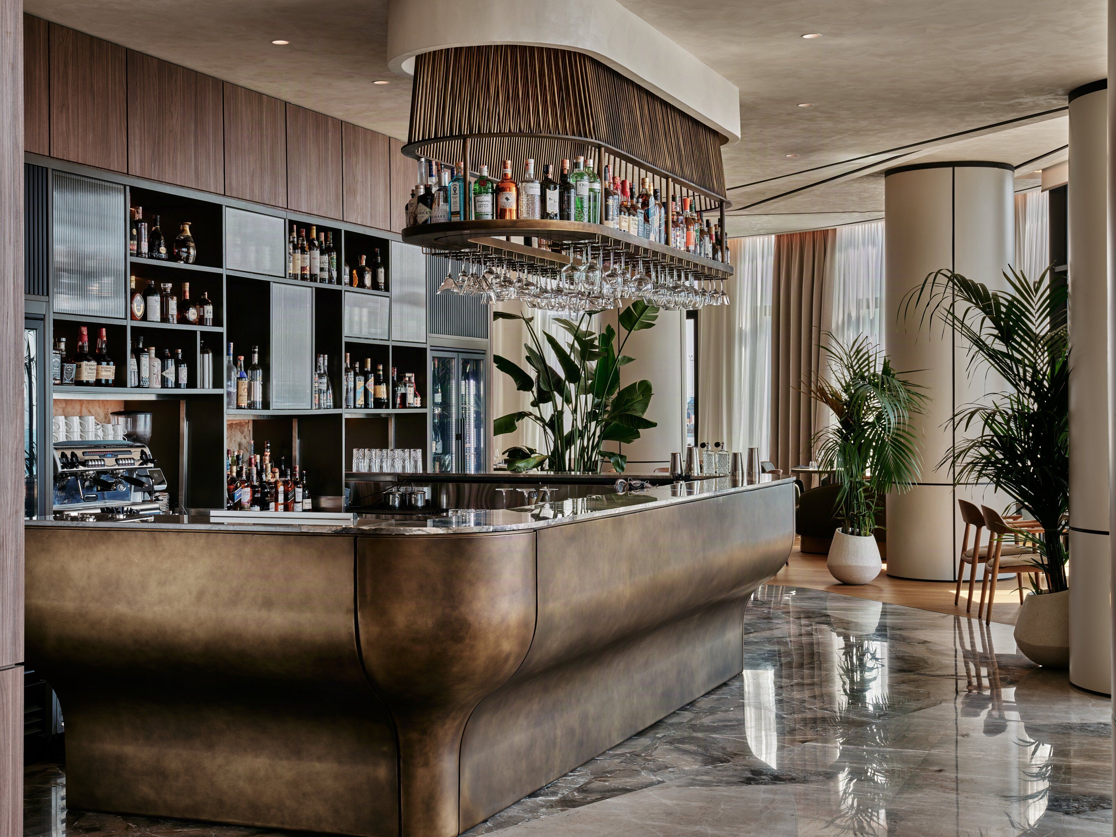 A modern bar area with a curved counter, shelves of liquor bottles, a coffee machine, and a large potted plant.