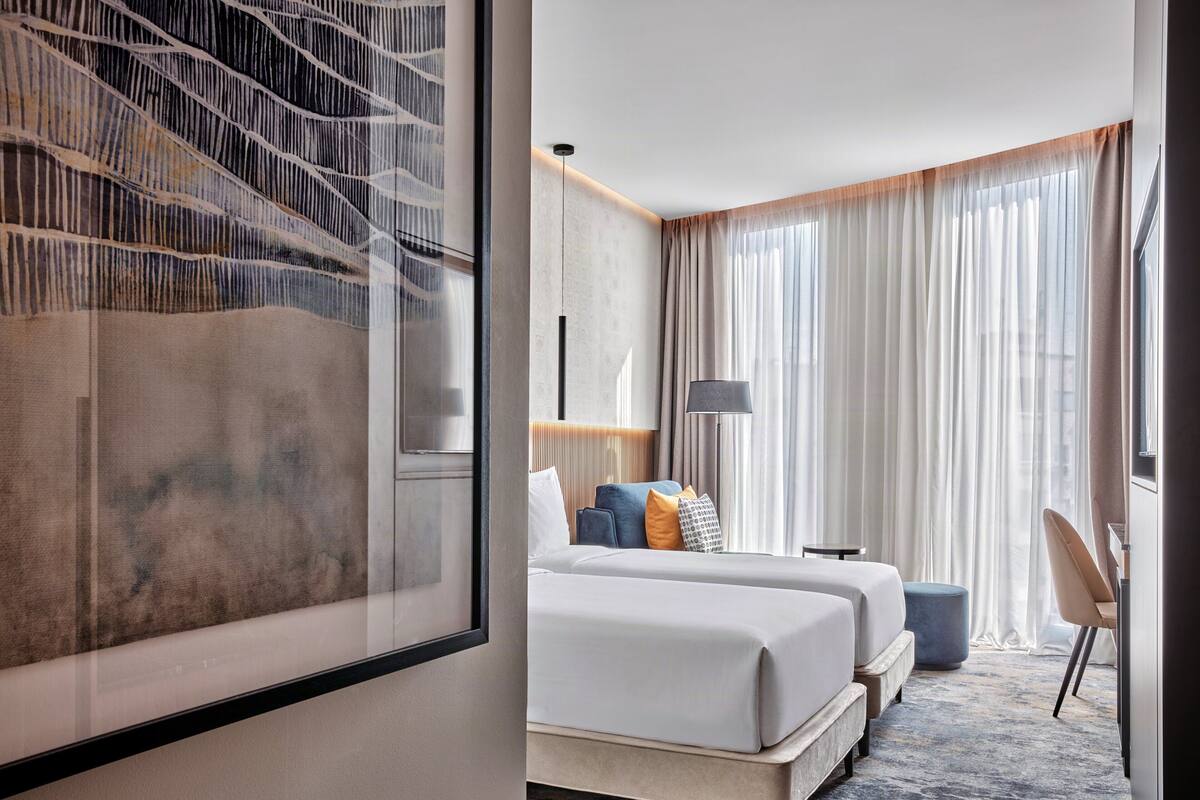Ein Hotelzimmer mit einem Bett, zwei Nachttischen, einer Lampe, einem Sessel und einem großen Fenster mit Vorhängen.