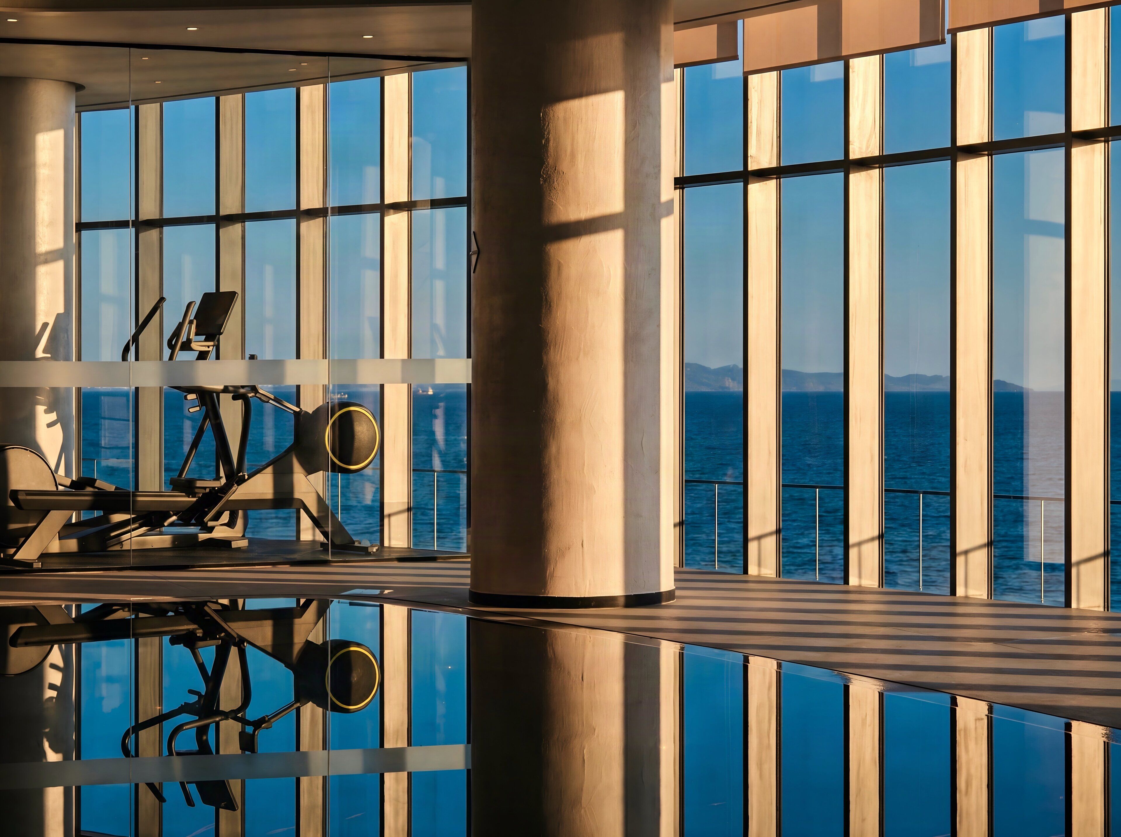 A gym with exercise equipment and large windows overlooking the sea.