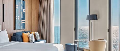 A modern hotel room with a large bed, a seating area with a sofa and ottoman, a standing lamp, and a large window offering a view of the sea.