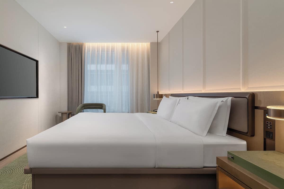Habitación de hotel moderna con una cama grande, un televisor de pantalla plana en la pared, una mesita de noche con lámpara y vistas a la ciudad a través de cortinas translúcidas.