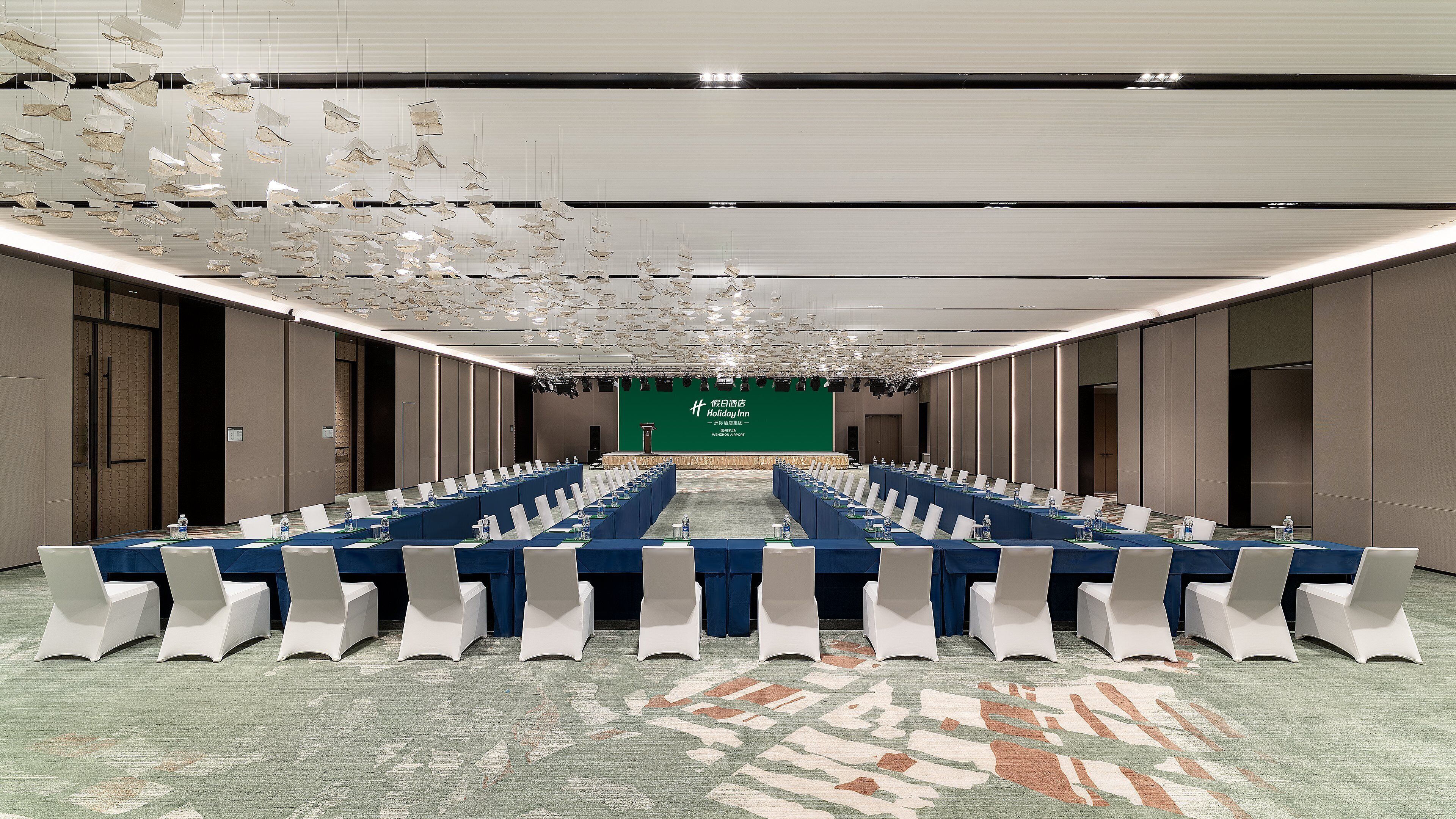 Un salón de conferencias con una larga mesa, sillas blancas y una pantalla verde al frente.