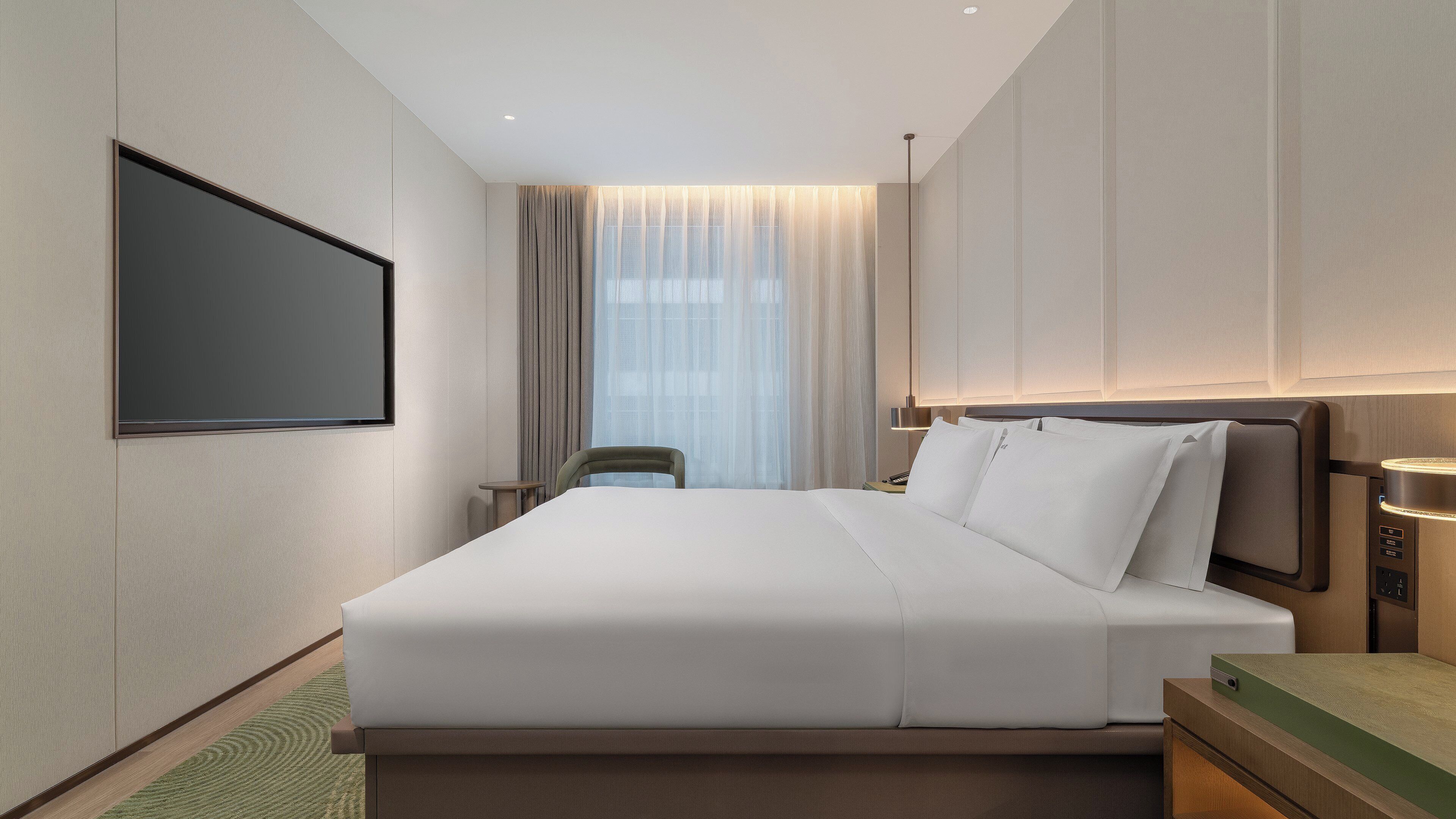 Ein modernes Hotelzimmer mit einem großen Bett, einem flachbildfernseher an der Wand, einem Nachttisch mit Lampe und Blick auf die Stadt durch transparente Vorhänge.