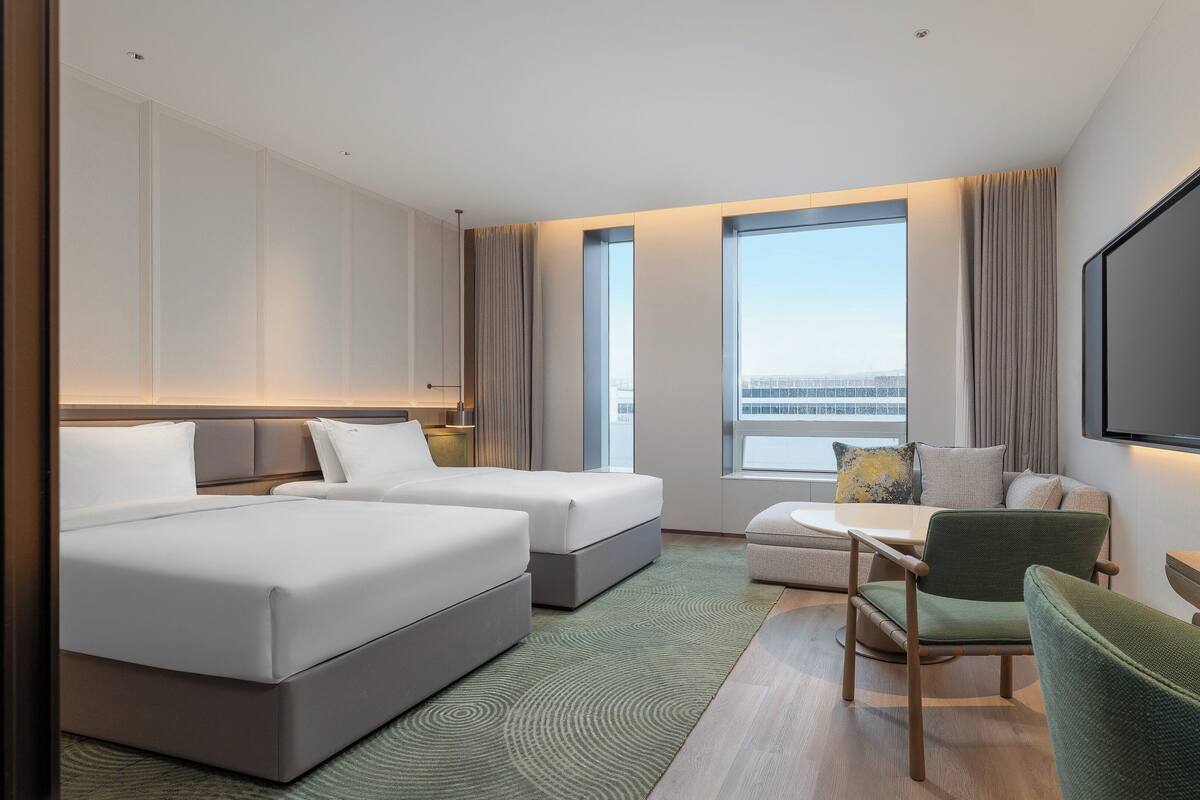 Habitación de hotel con cama, dos mesitas de noche, sofá, sillón, televisor y ventanas amplias con vistas.