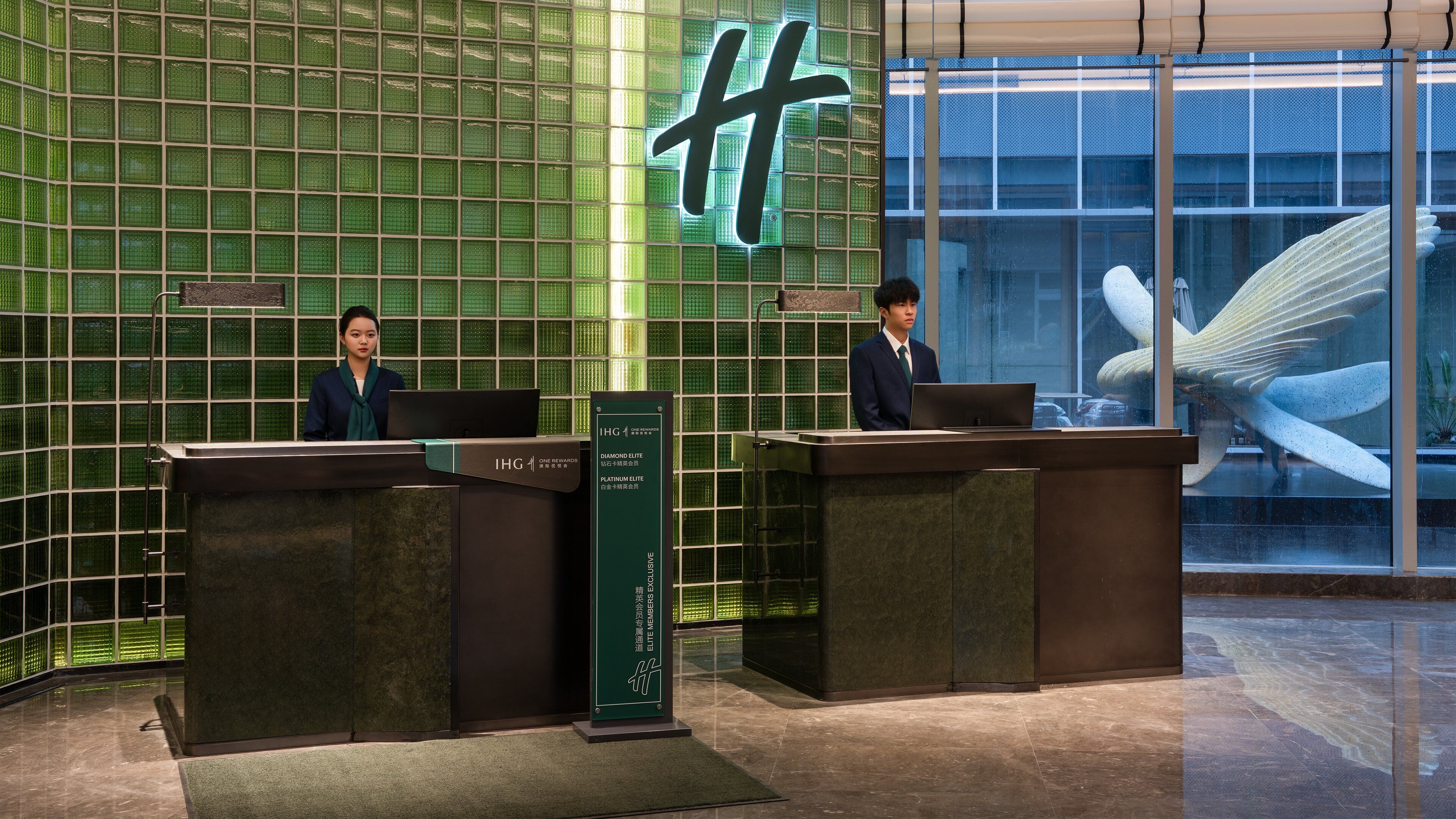 A reception area with a green mosaic wall, two receptionists, and a large glass window with a sculpture.
