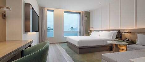 A modern hotel room with a large bed, a desk, a green chair, a small table, and a view of a building through the window.