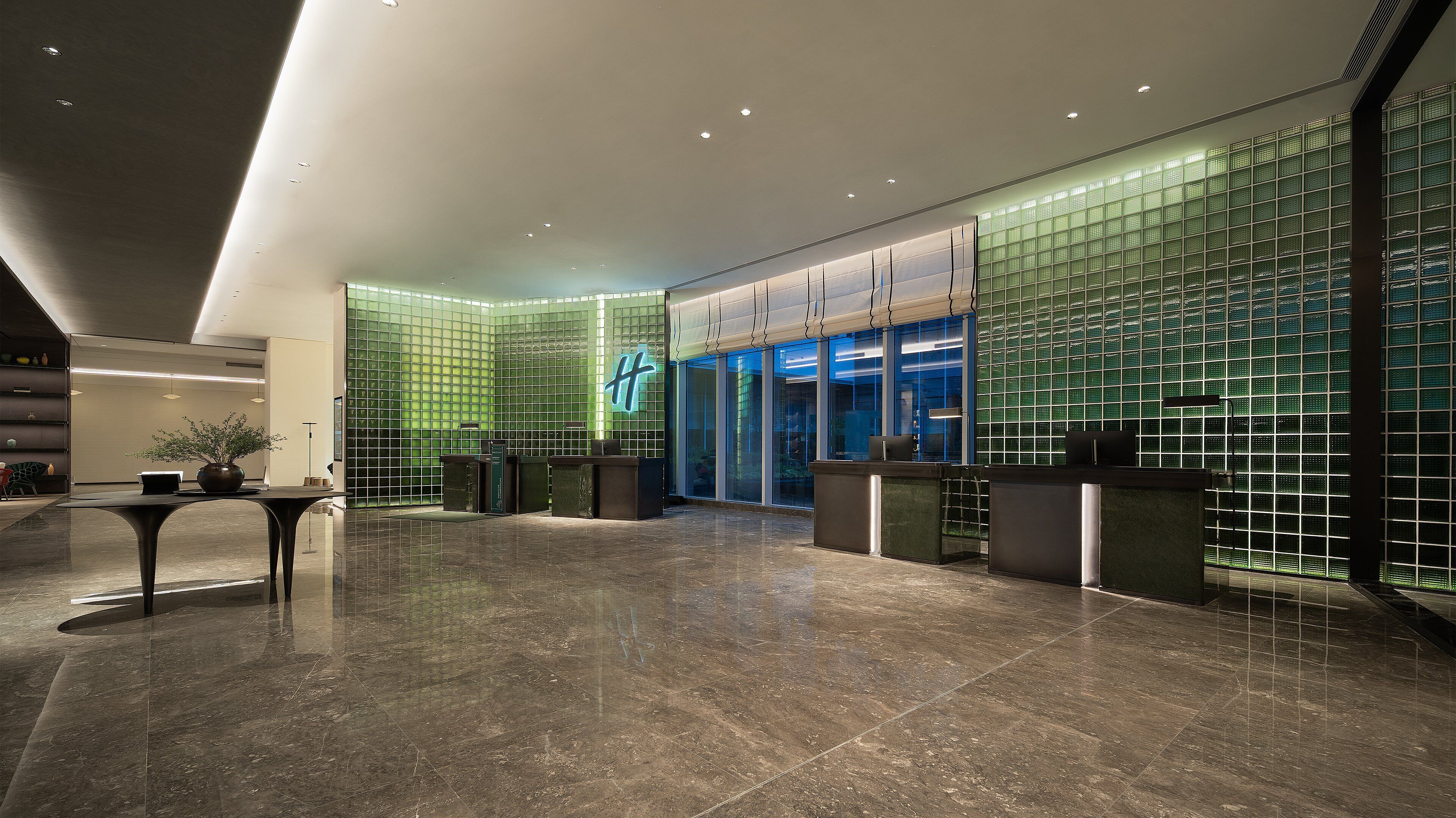 A modern hotel lobby with a reception desk, a potted plant, and a large glass door leading to the entrance.