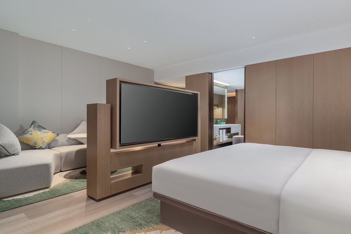 Una habitación de hotel moderna con una cama grande, un televisor de pantalla plana montado en un soporte de madera, una zona de estar con un sofá y sillones, y una mesita pequeña.