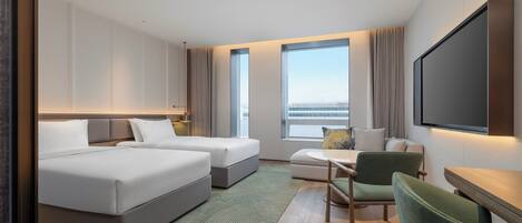A hotel room with a bed, two bedside tables, a sofa, a chair, a TV, and large windows with a view.